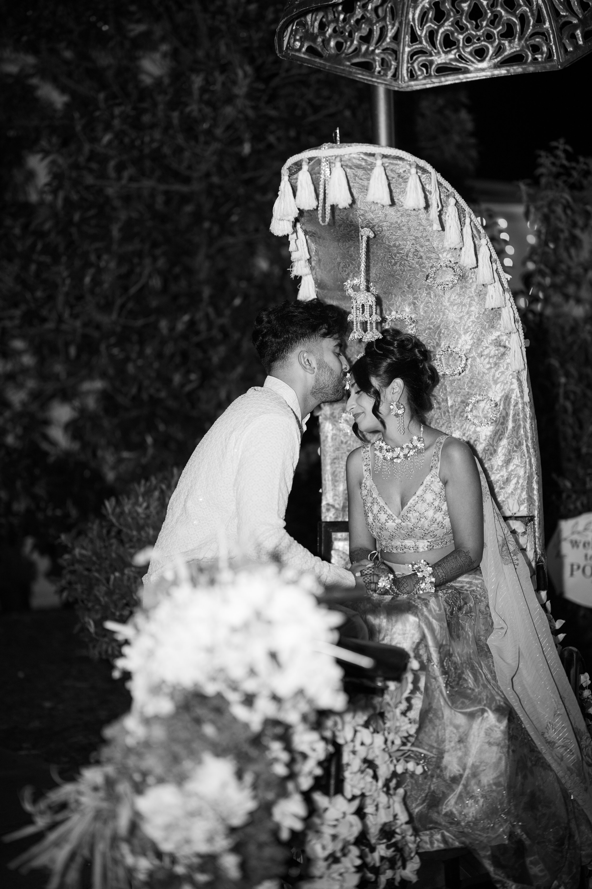 A black and white photo of a couple leaning their foreheads together during a wedding ceremony, with the groom wearing traditional Indian attire and the bride dressed in an ornate outfit with jewelry and henna on her hands, seated underneath a decora