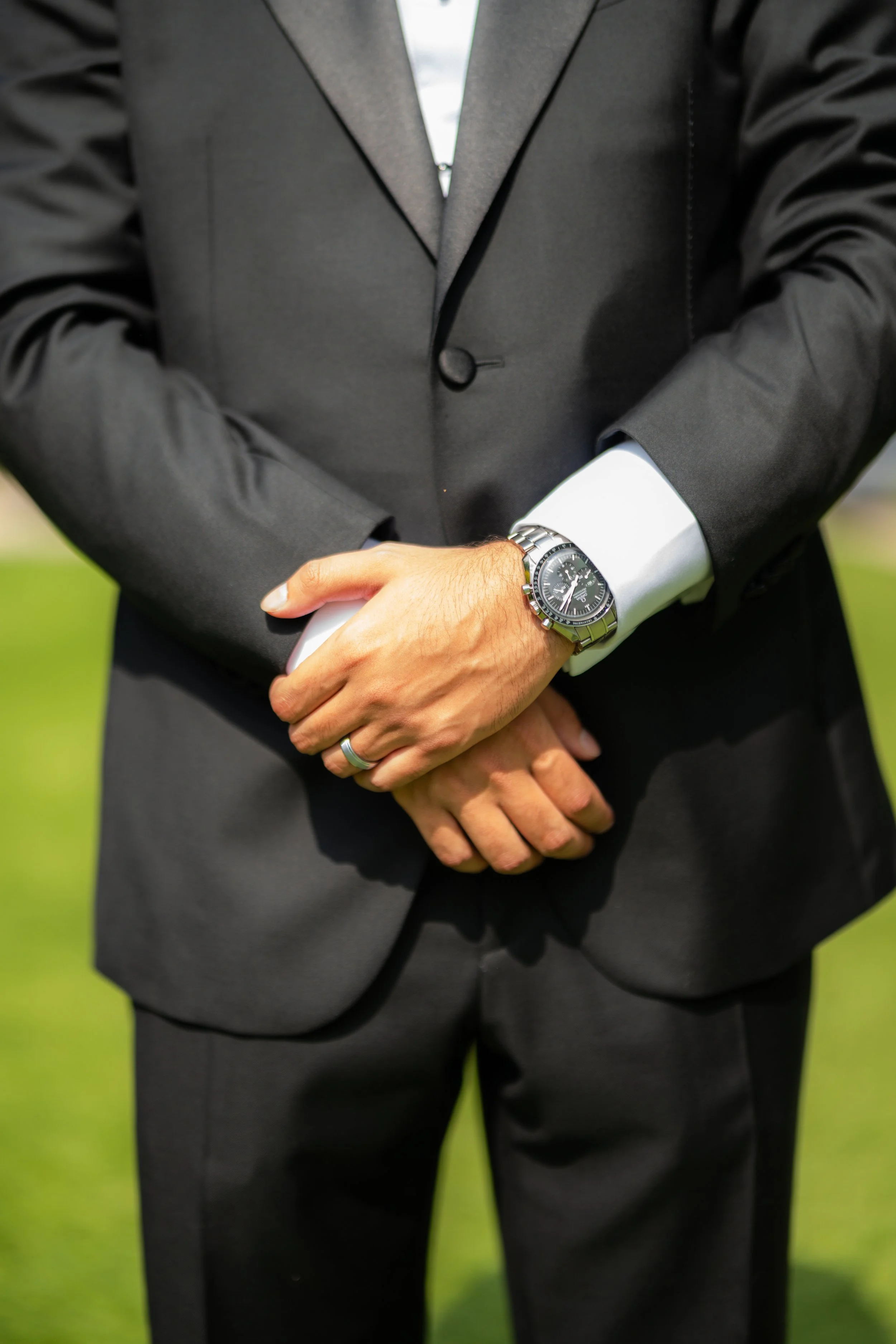 Close-up of a man in a black suit with a white shirt, standing with arms crossed, wearing a wristwatch, outdoors with a blurred green background.