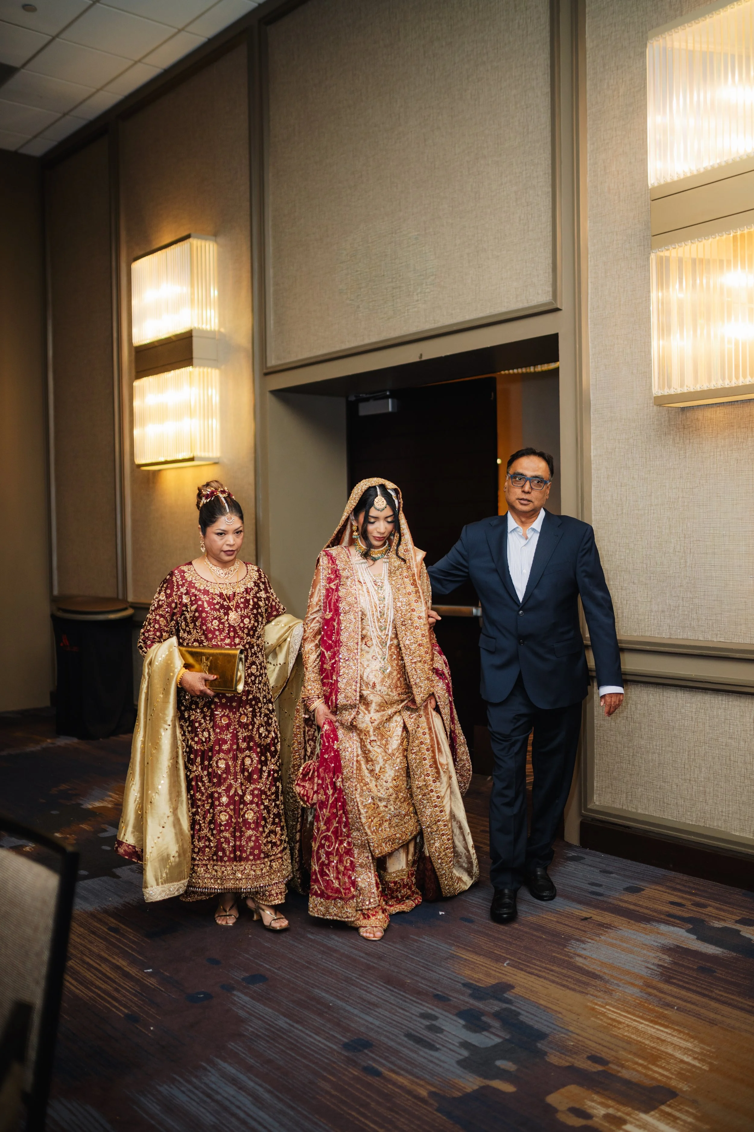 Three people, two women in traditional Indian attire and one man in a suit, walking together in a hotel corridor.