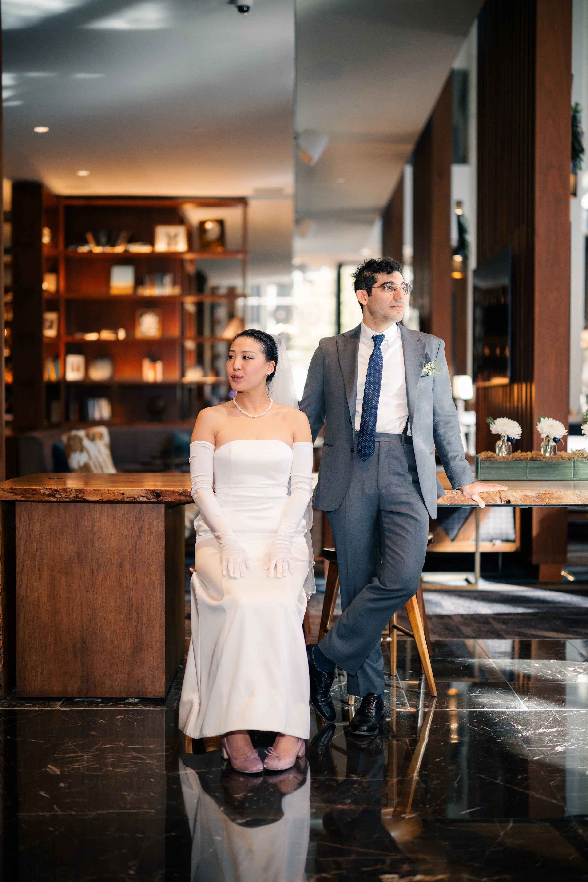 A bride and groom are in a modern interior setting, with the bride seated and wearing a strapless white wedding gown and long gloves, and the groom standing next to her in a gray suit with a blue tie, holding a hand on the table and looking to the side.