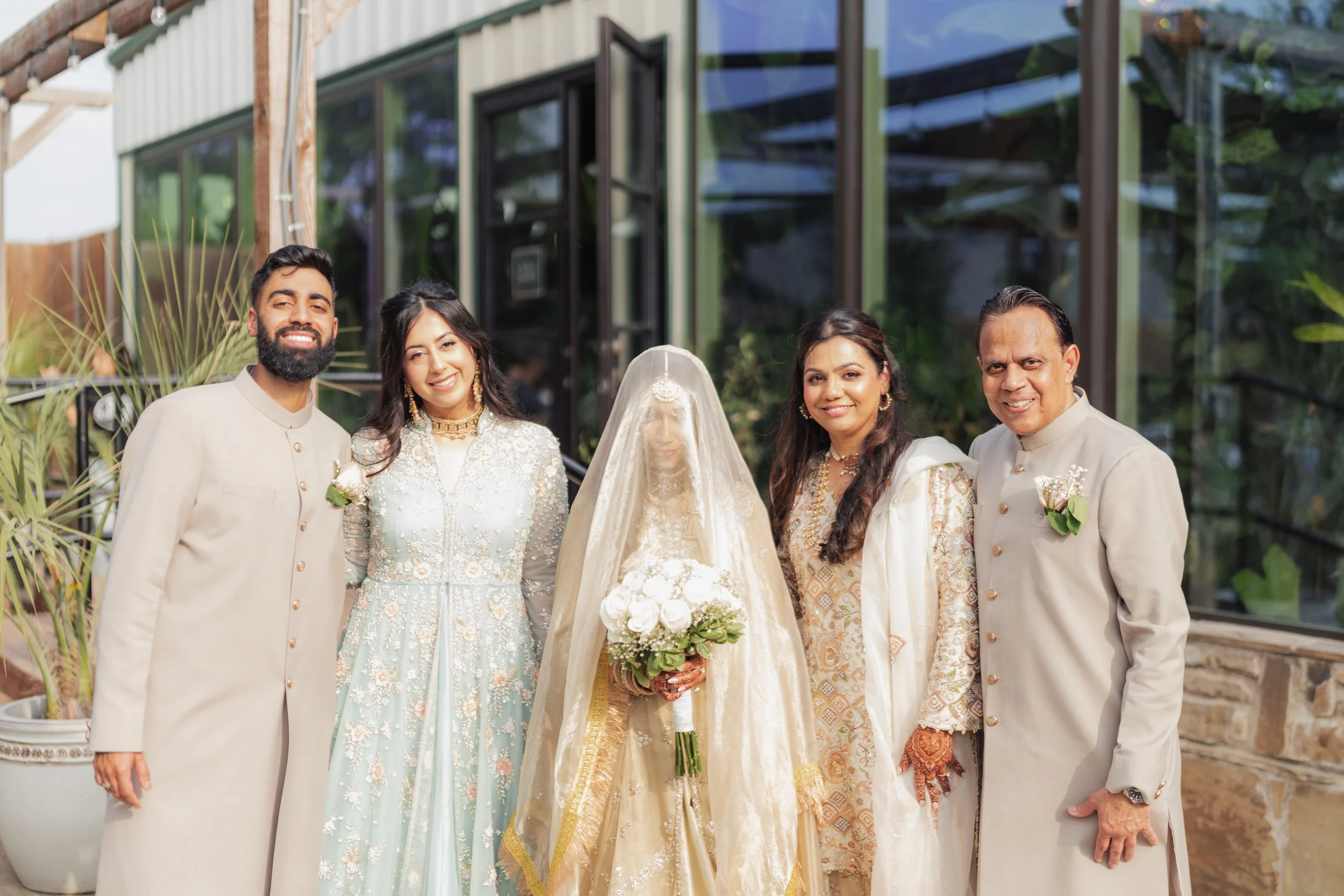 Group of five people dressed in traditional South Asian wedding attire, standing outside in front of a glass building and greenery. The bride is wearing a gold and cream outfit with a veil, holding a bouquet, and flanked by three women and one man.
