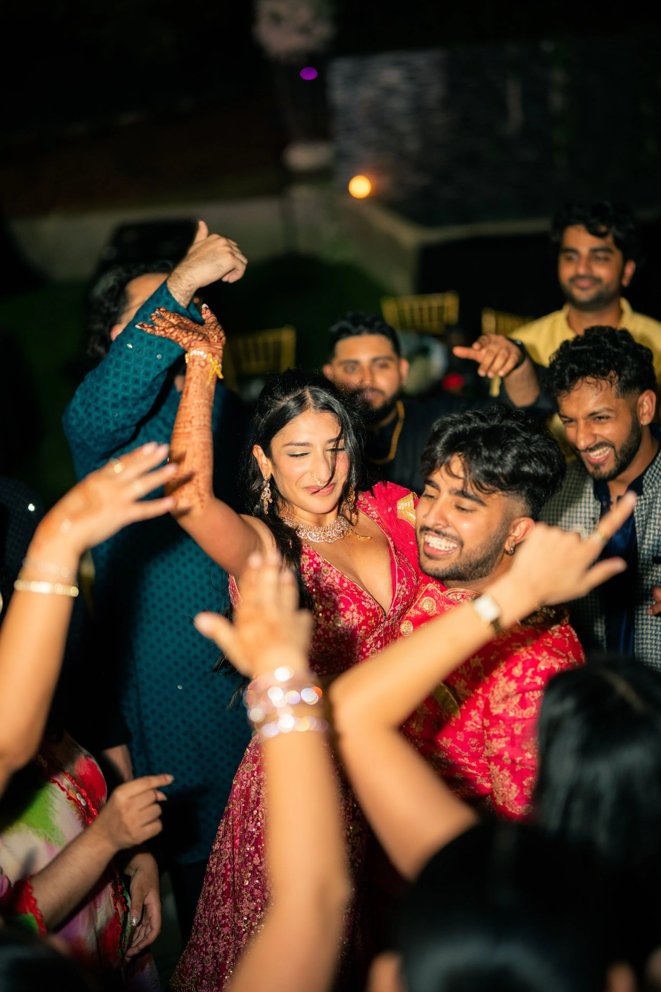 Group of people celebrating at a party, dancing closely together outdoors at night, with smiling faces and colorful traditional attire.