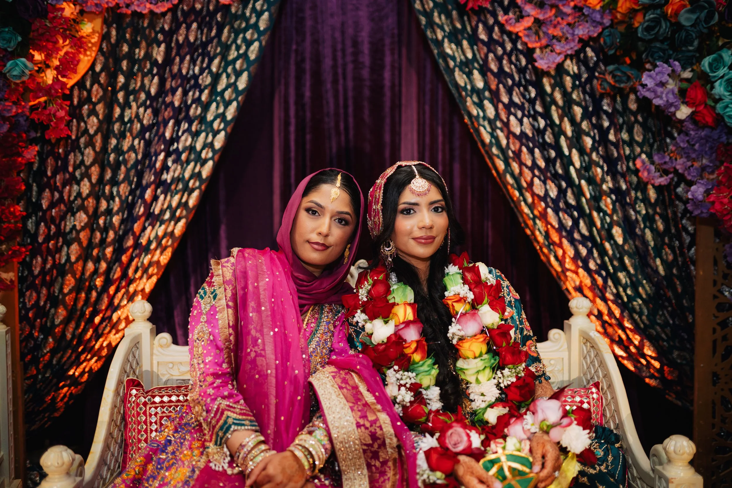 Two women dressed in traditional Indian attire sitting on a decorated wedding stage with a floral backdrop, holding bouquets of flowers.