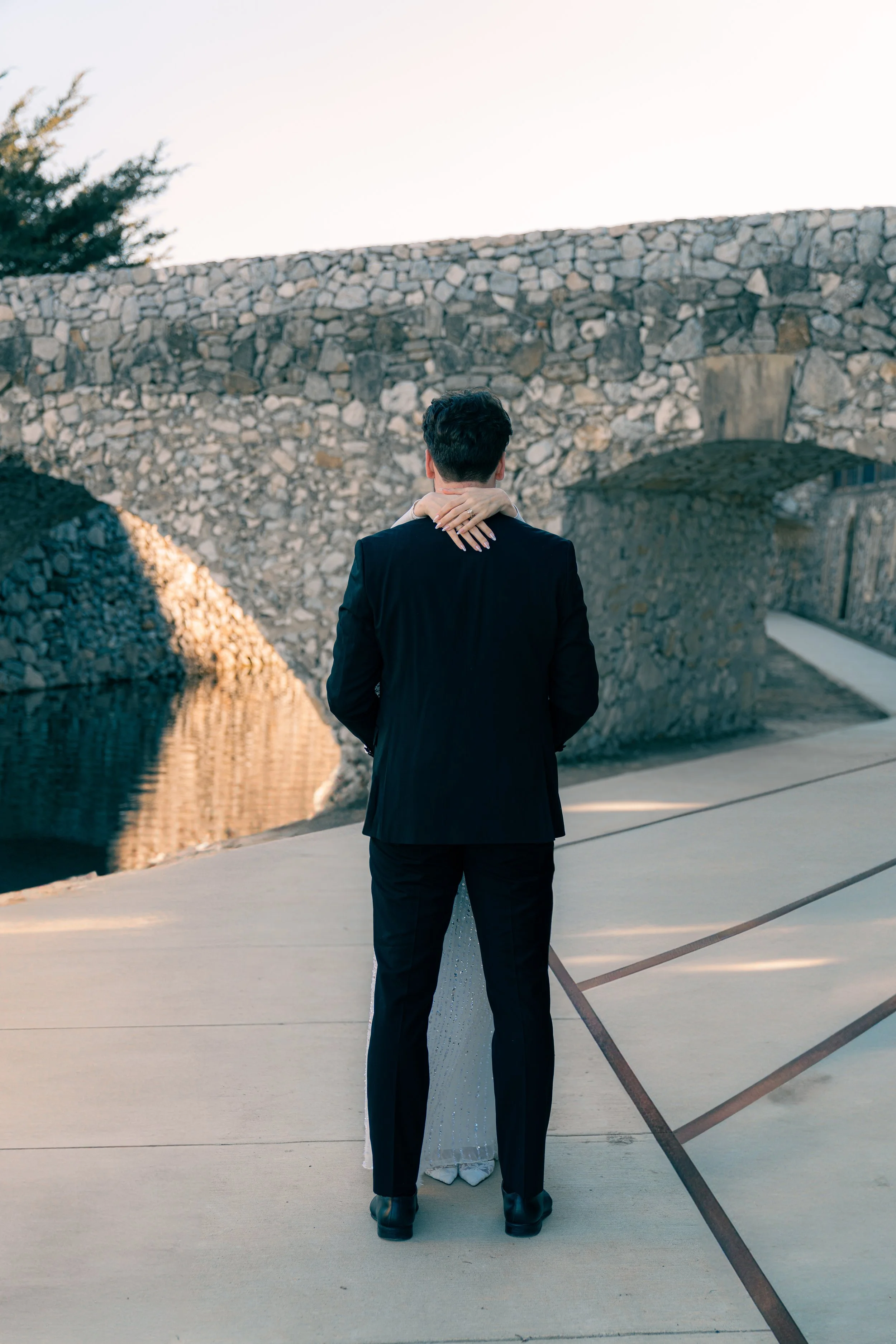 A man in a black suit standing on a concrete pathway by a stone bridge, embracing a woman whose face is not visible, with water reflecting the bridge and a clear sky in the background.