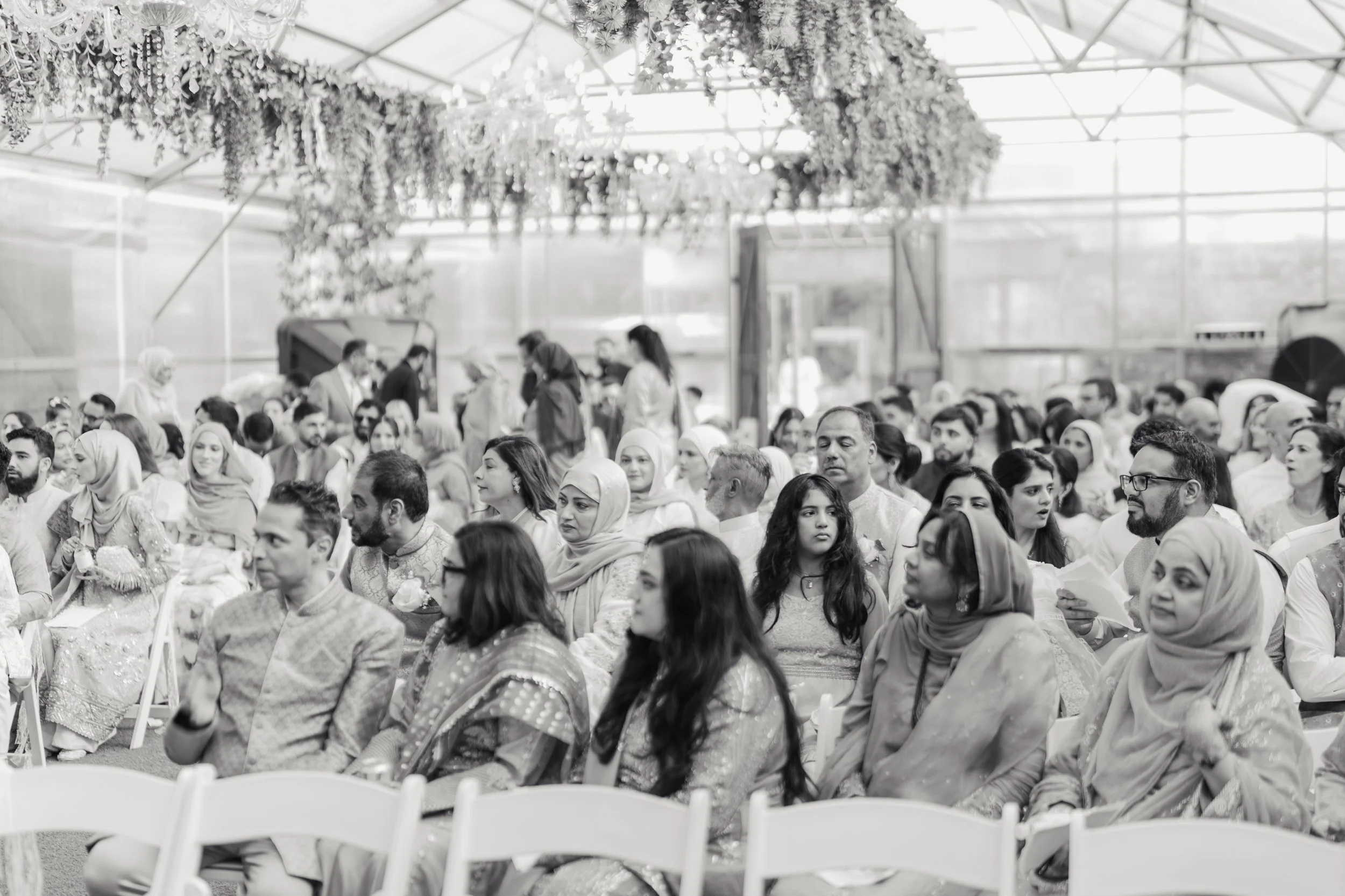 A large group of people seated inside a greenhouse decorated with hanging flowers and greenery. Many women are wearing headscarves, and everyone appears to be attending a formal event or gathering.