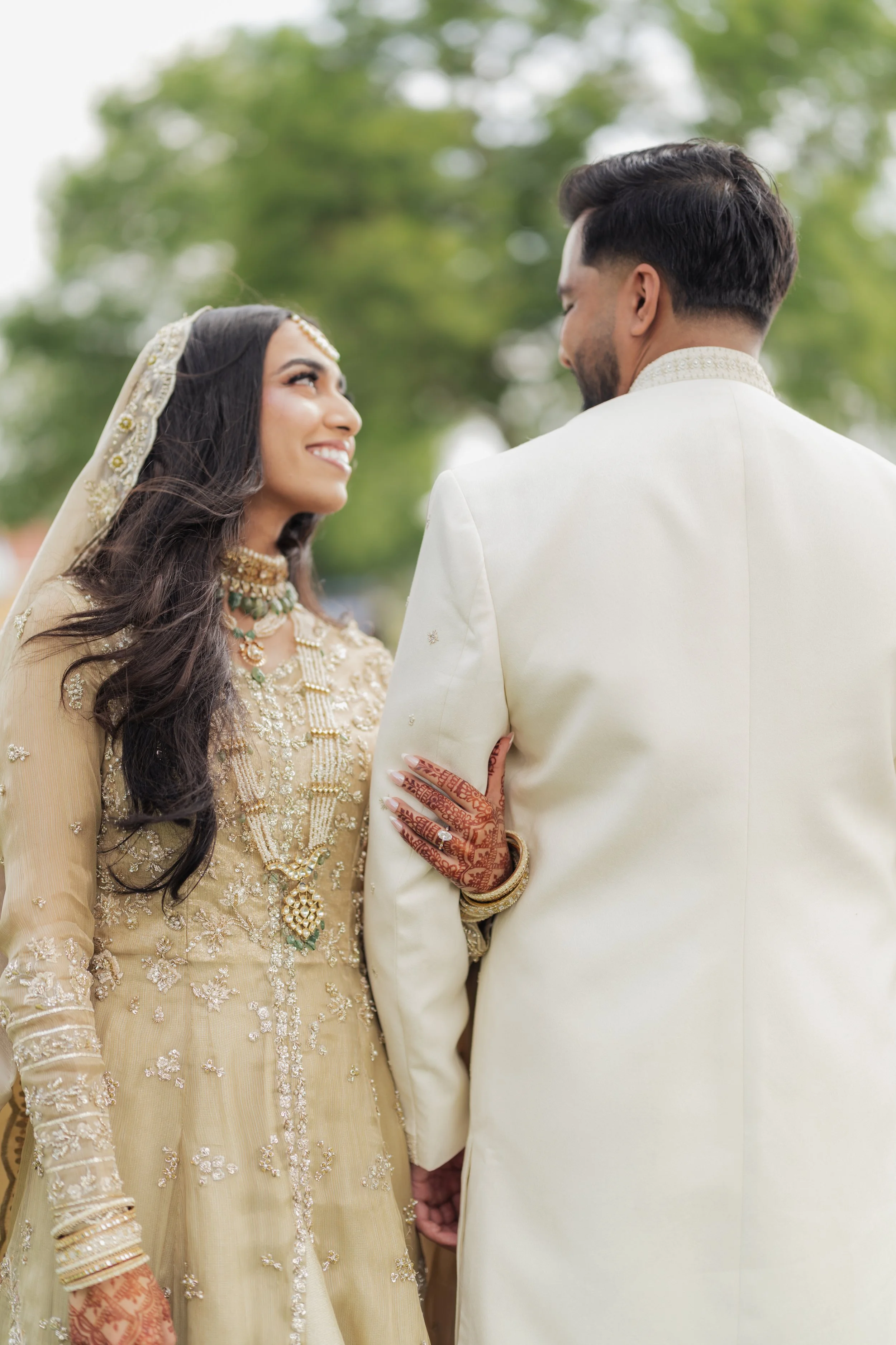 A bride and groom holding hands during their wedding ceremony, with the bride dressed in an ornate gold traditional outfit and the groom in a cream-colored suit, outdoors with green trees in the background.