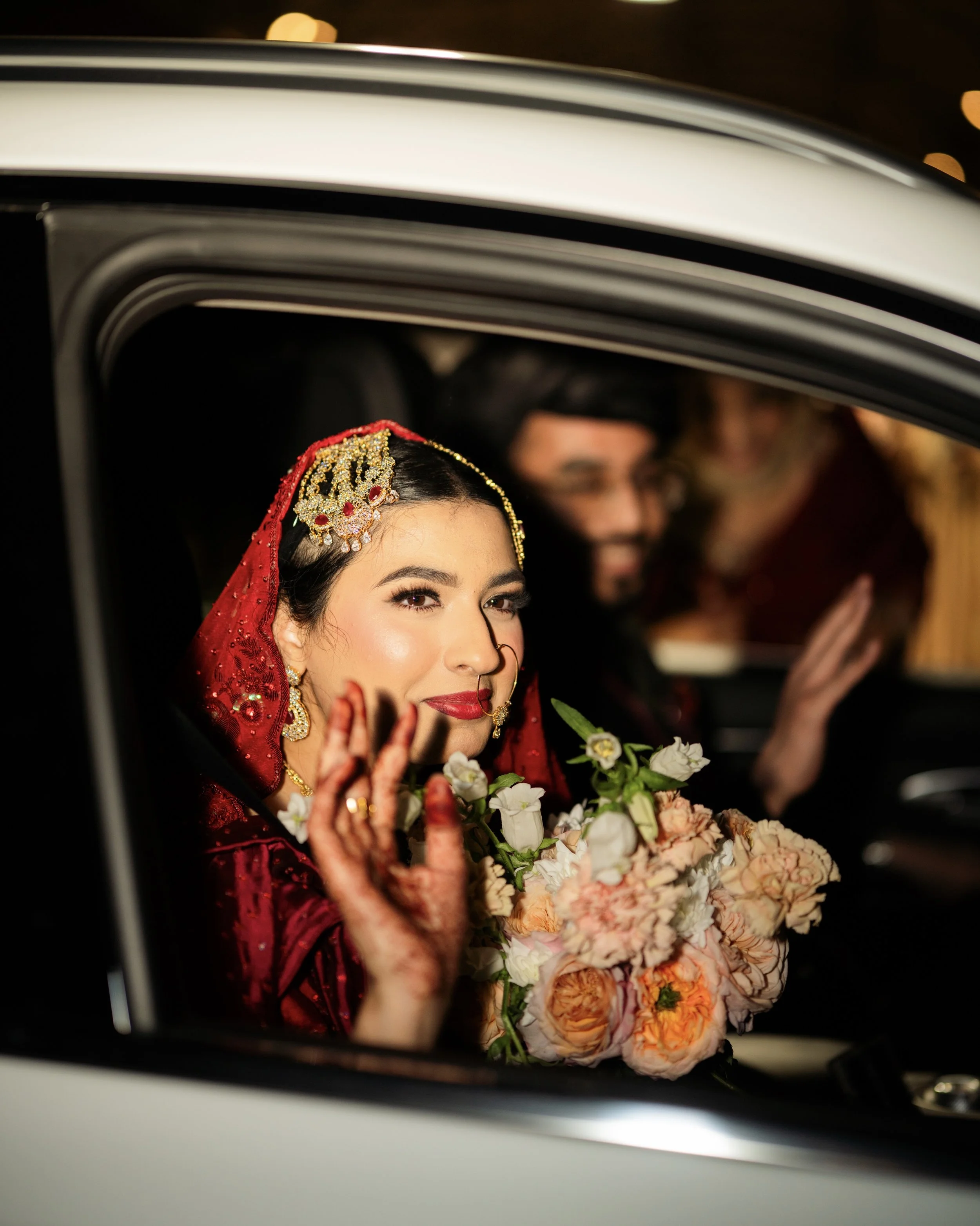 Close-up of a bride in a red traditional wedding dress and jewelry, sitting in a car, holding a bouquet of flowers, and waving.