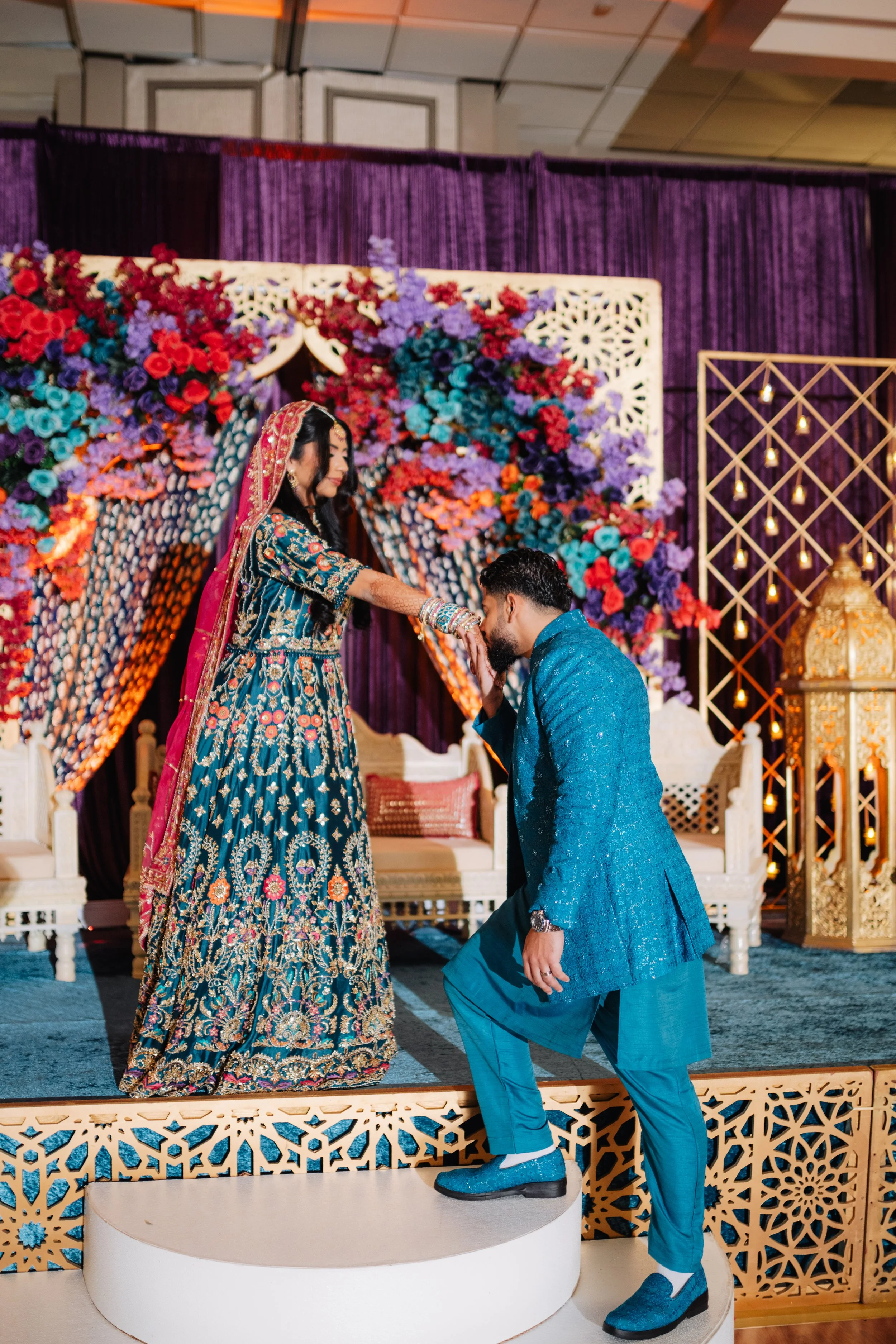 A couple at a wedding ceremony. The woman wearing a dark blue traditional dress with colorful embroidery and a head covering, and the man in a blue traditional suit. The man is kneeling and kissing the woman's hand. The background is decorated with c