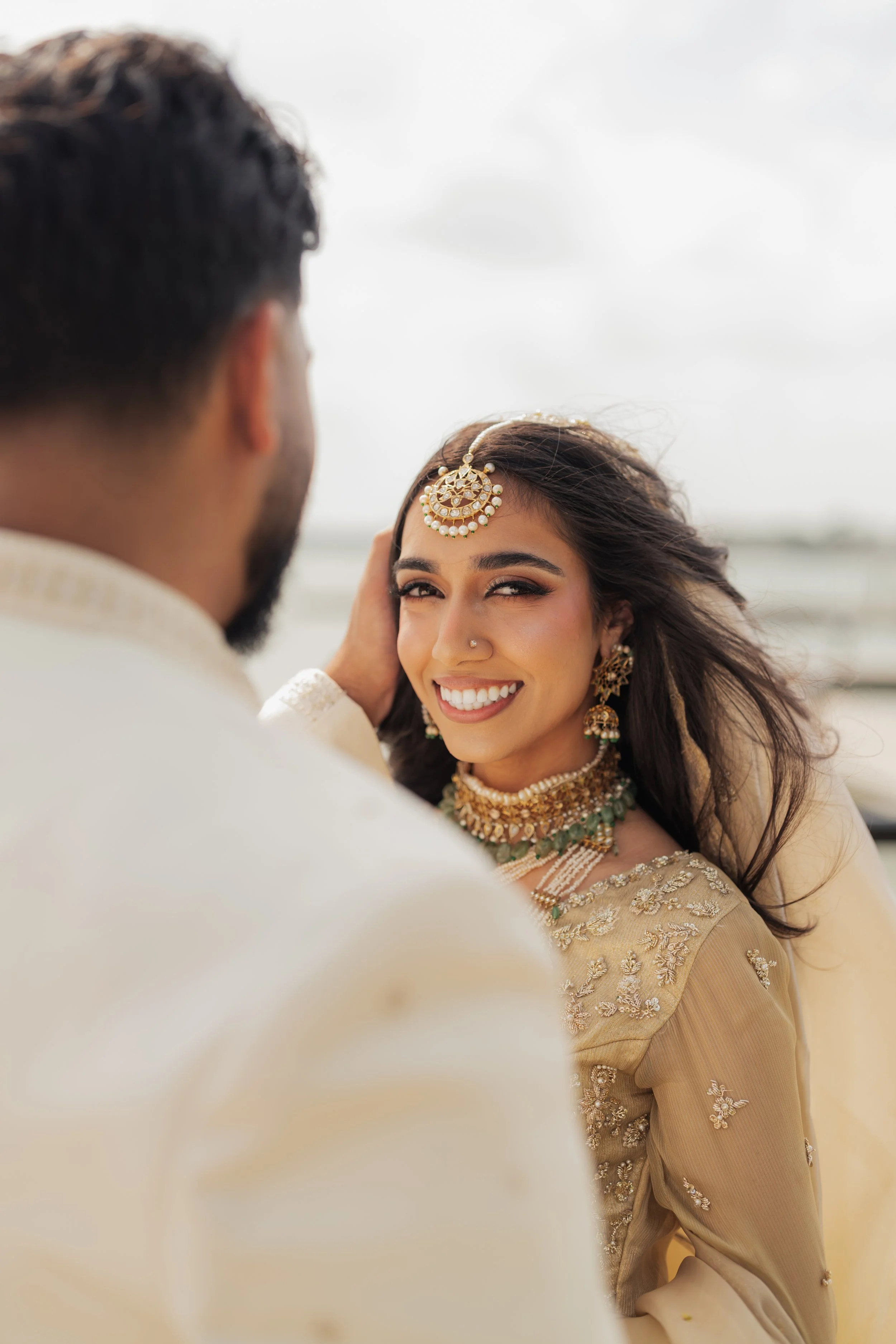 A woman wearing traditional Indian jewelry and attire smiling, with a man touching her face, on the beach with cloudy sky in the background.