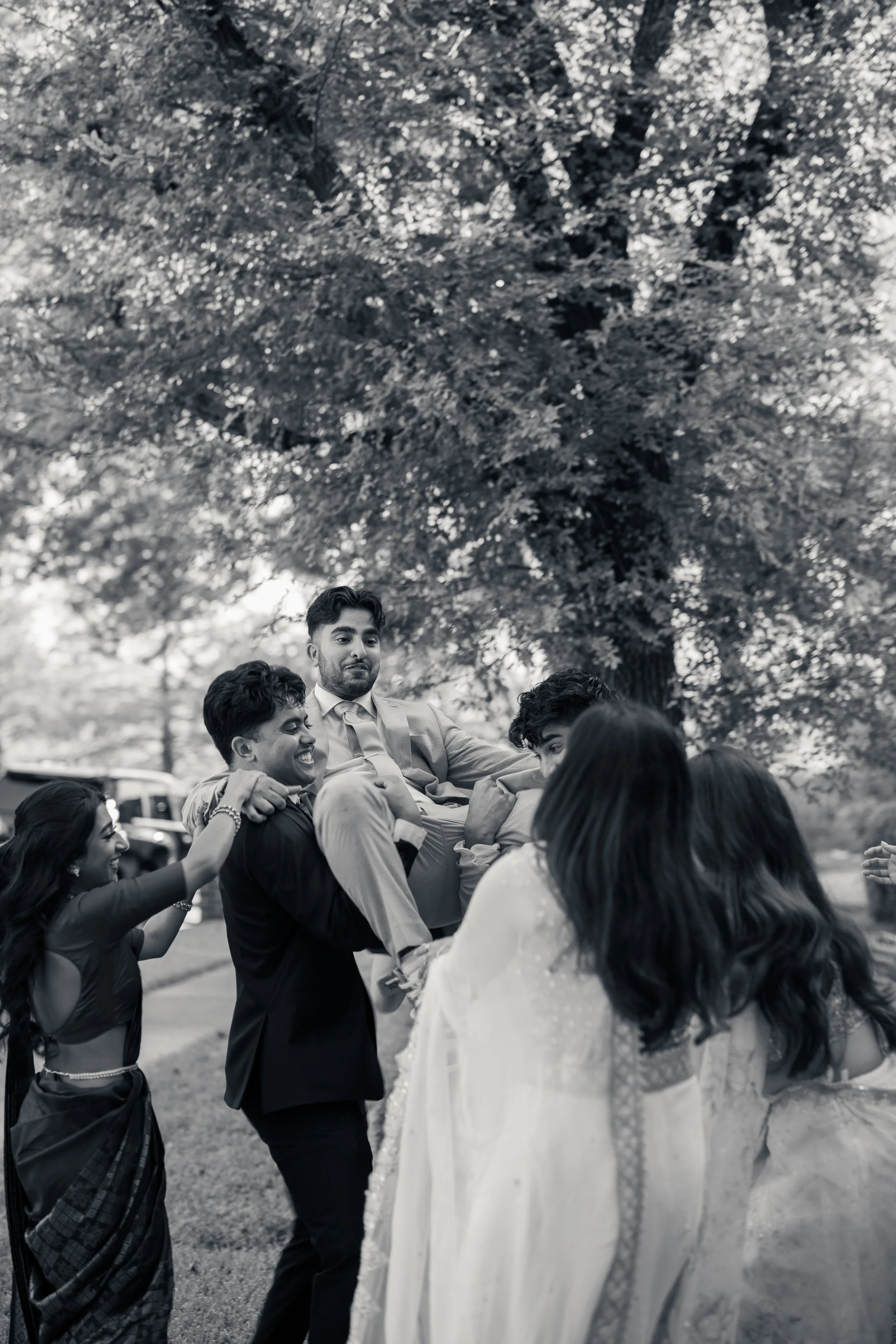 Group of people celebrating outdoors under a large tree, lifting a man in a suit, with women in traditional attire smiling and cheering.