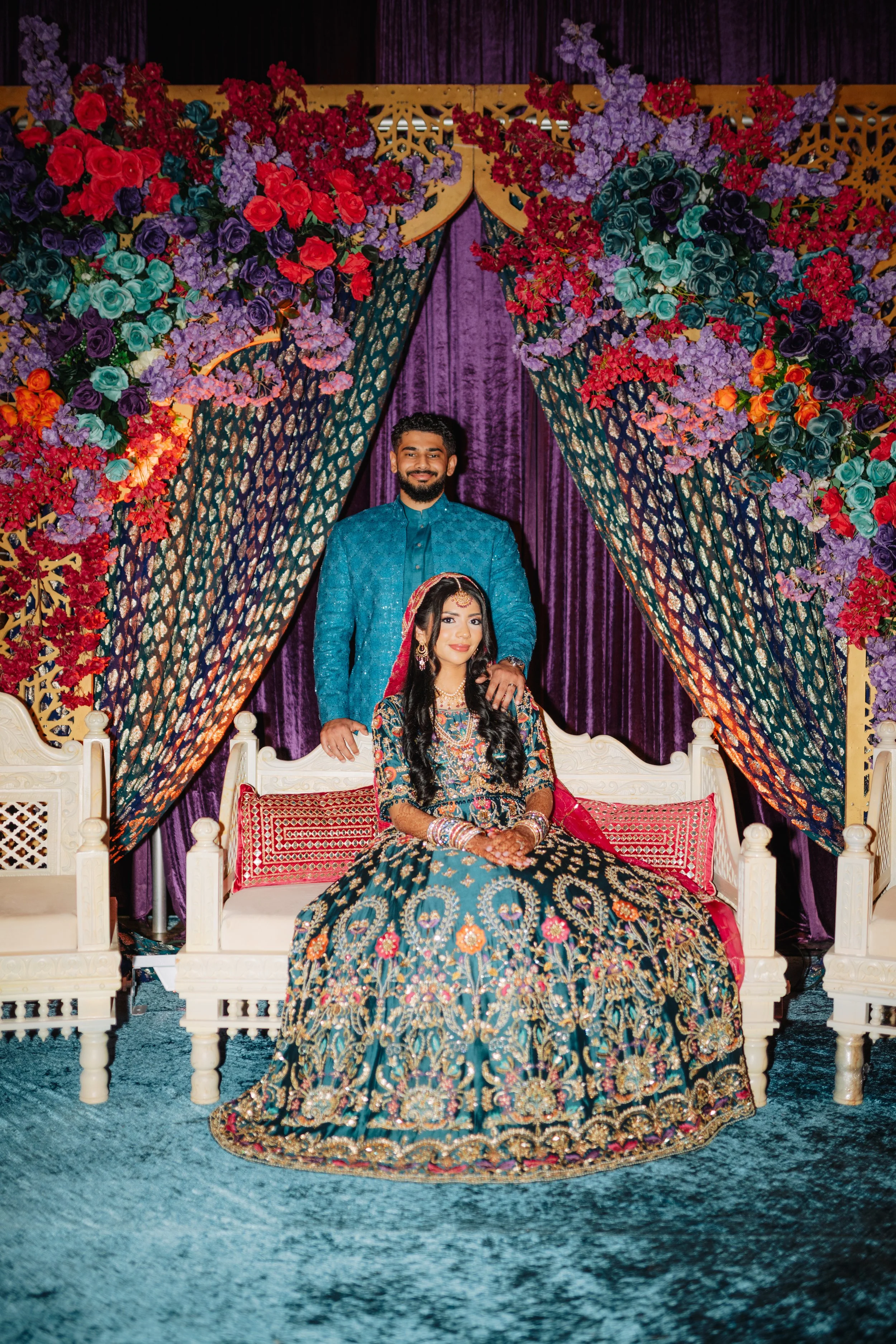 A couple in colorful traditional Indian wedding attire sitting and standing on a white ornate bench beneath a floral canopy of purple, red, and pink flowers, with purple curtains in the background.