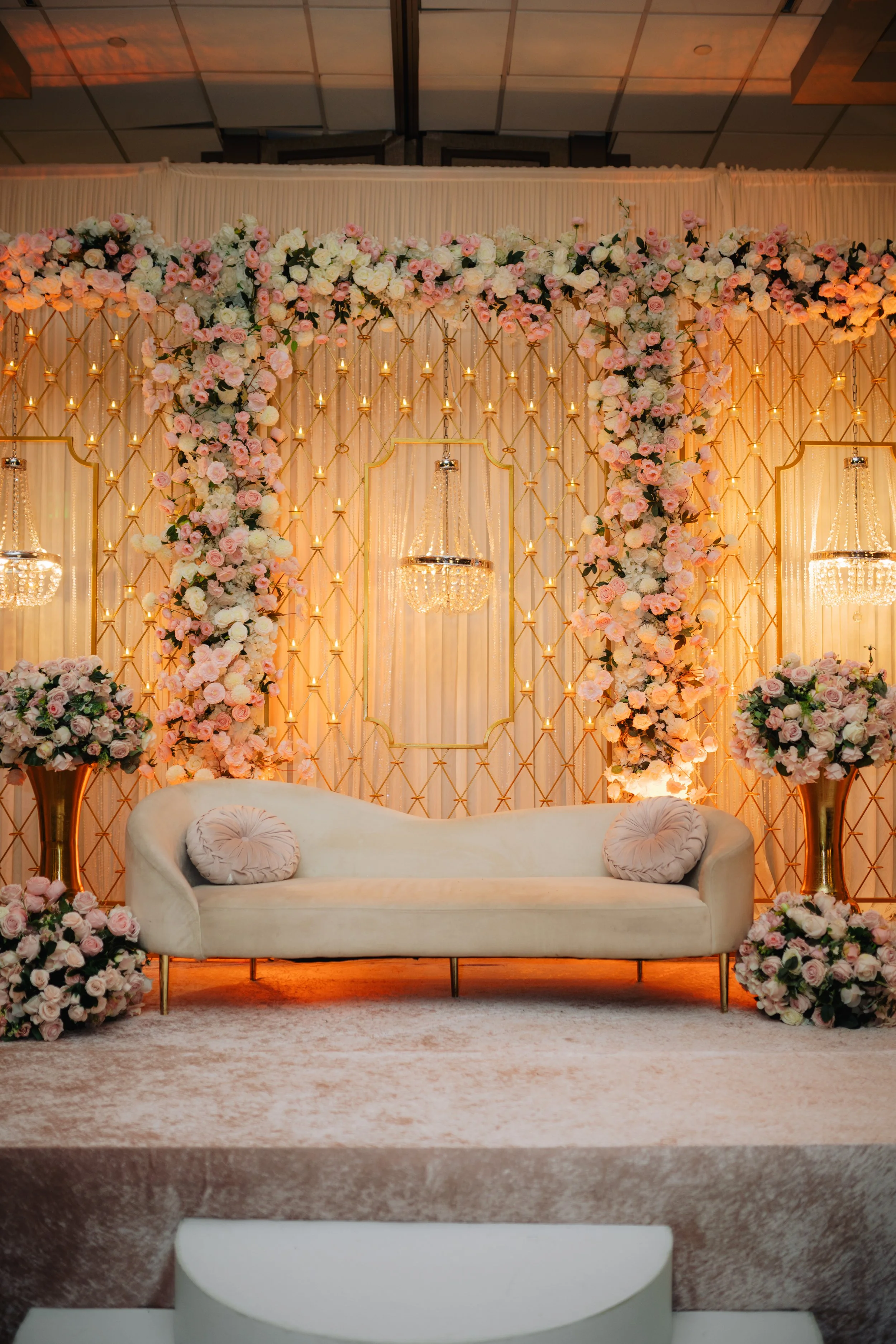 Elegant indoor wedding backdrop decorated with an abundance of pink and white roses, gold geometric accents, and chandelier lighting. A white plush sofa with matching pillows is positioned in front of the backdrop, set on a light-colored carpet.
