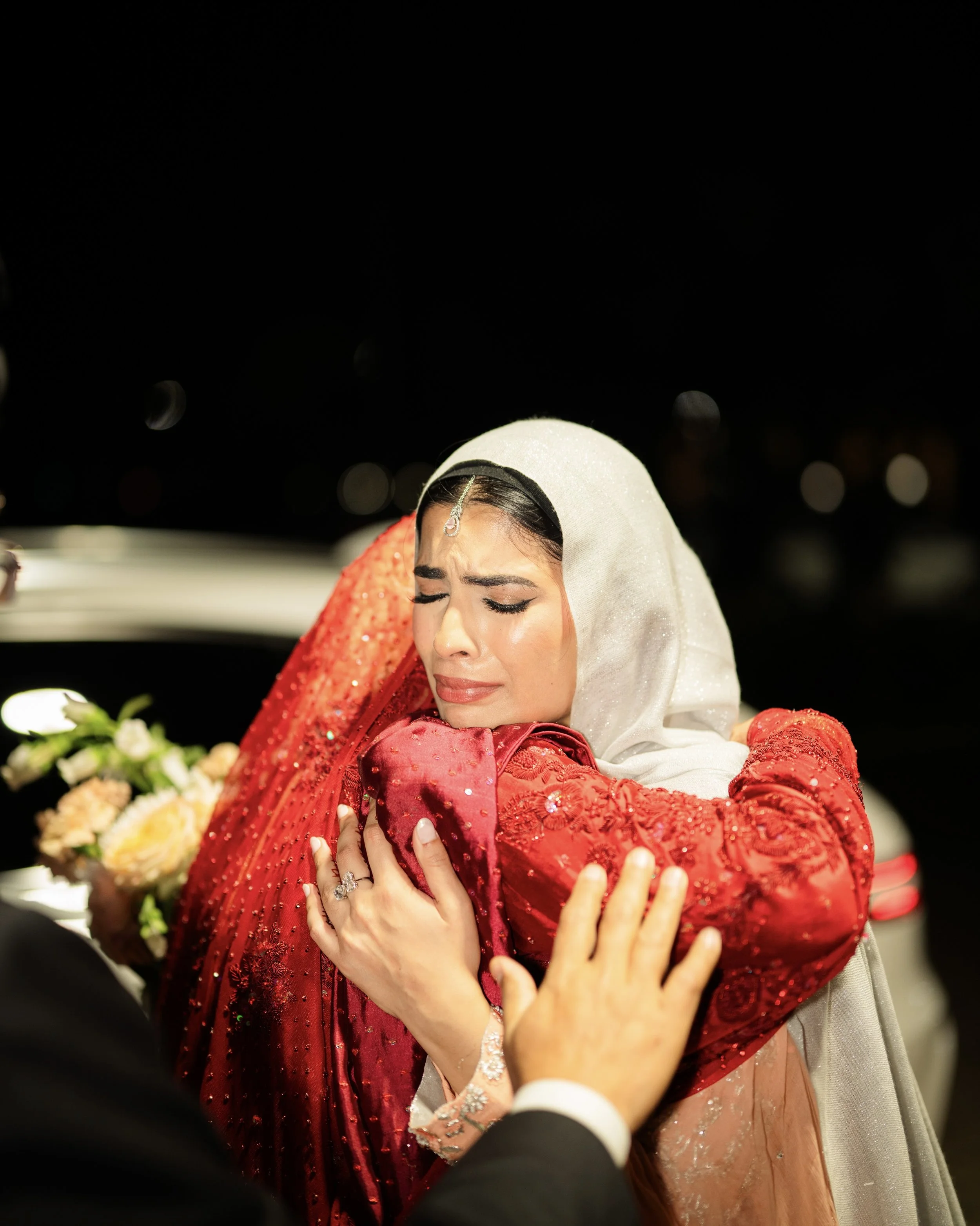 Two women hugging, one in a red dress and the other in a cream hijab, with emotional facial expressions, at night with blurred background and a person's hand reaching out.