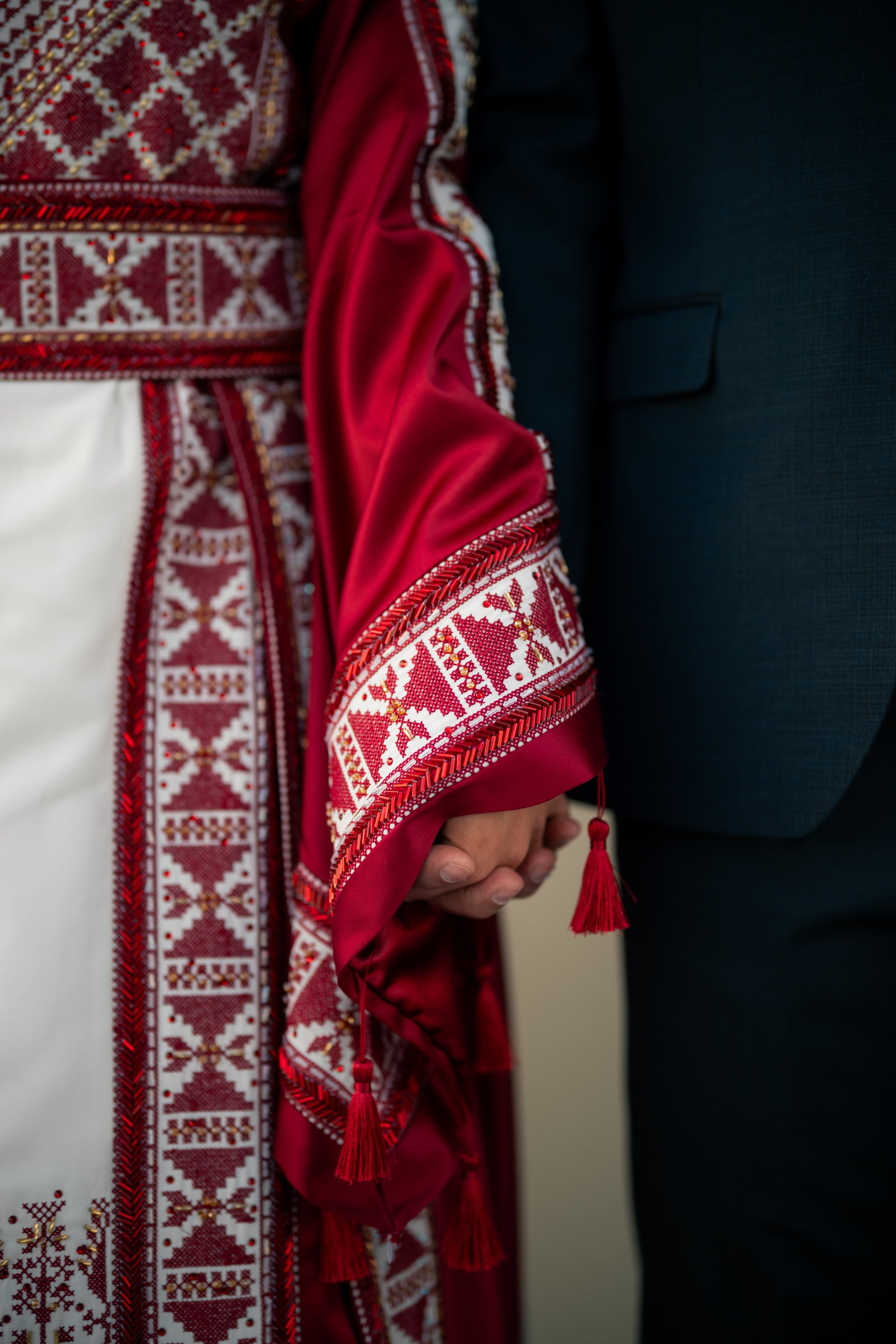 Close-up of two people holding hands, one person wearing a traditional embroidered garment with intricate red, white, and gold patterns and red tassels, the other person in a dark suit.