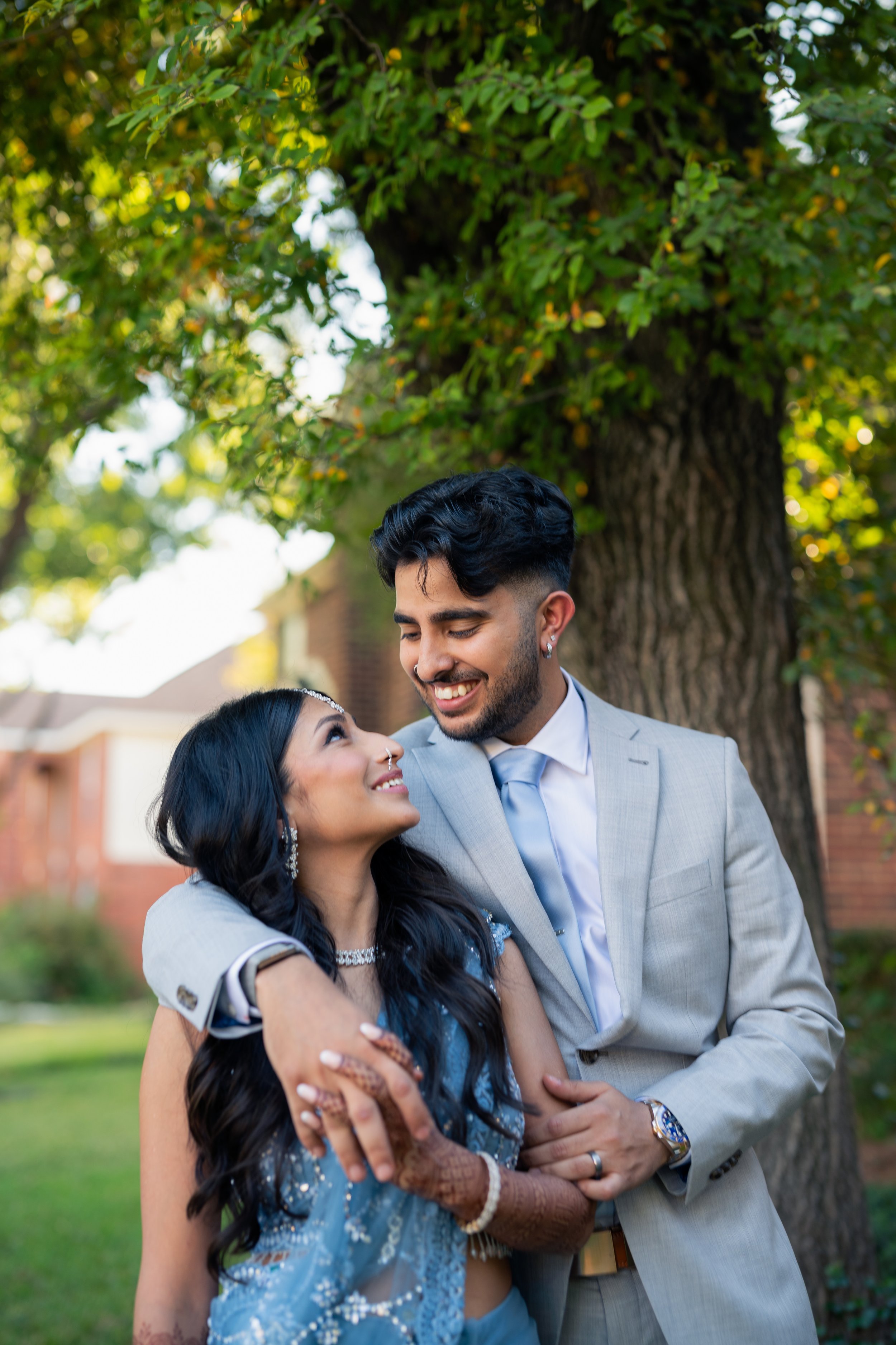 A couple dressed in formal wedding attire standing outdoors under a large tree, smiling and looking at each other affectionately.