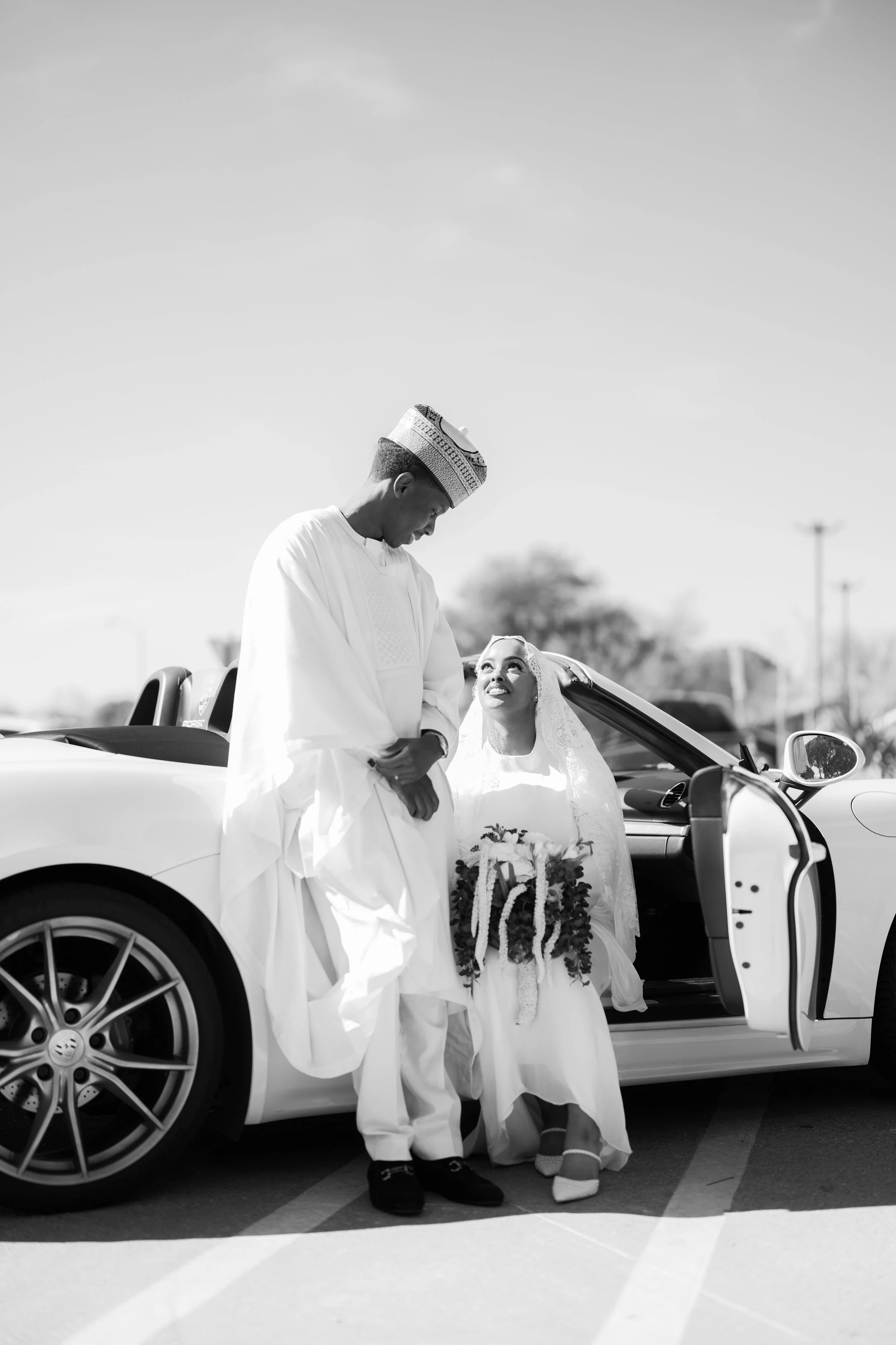 A black and white photo of a bride and groom on their wedding day, standing outside next to a convertible sports car. The bride is sitting in the car, holding a bouquet, looking up at the groom who is standing beside the car, smiling. The groom is we