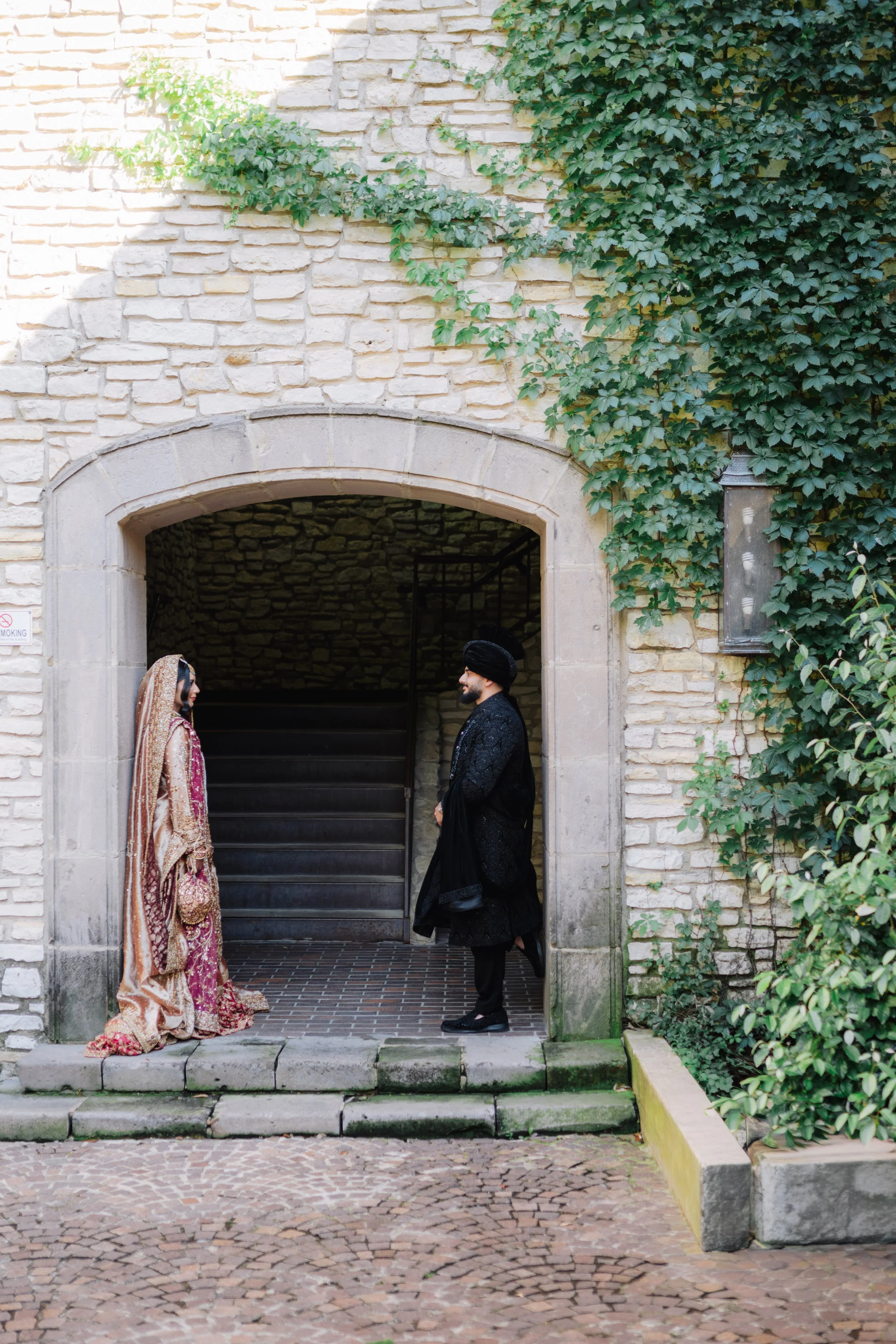 A woman dressed in traditional South Asian bridal attire and a man in formal South Asian clothing are standing face to face in front of an arched stone entrance adorned with green ivy on the wall.