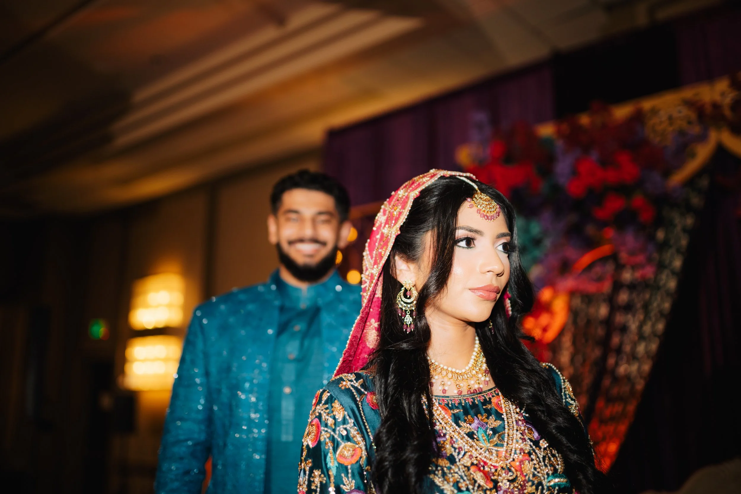 A bride and groom in traditional Indian wedding attire, standing indoors with a decorated backdrop of colorful flowers and fabrics.