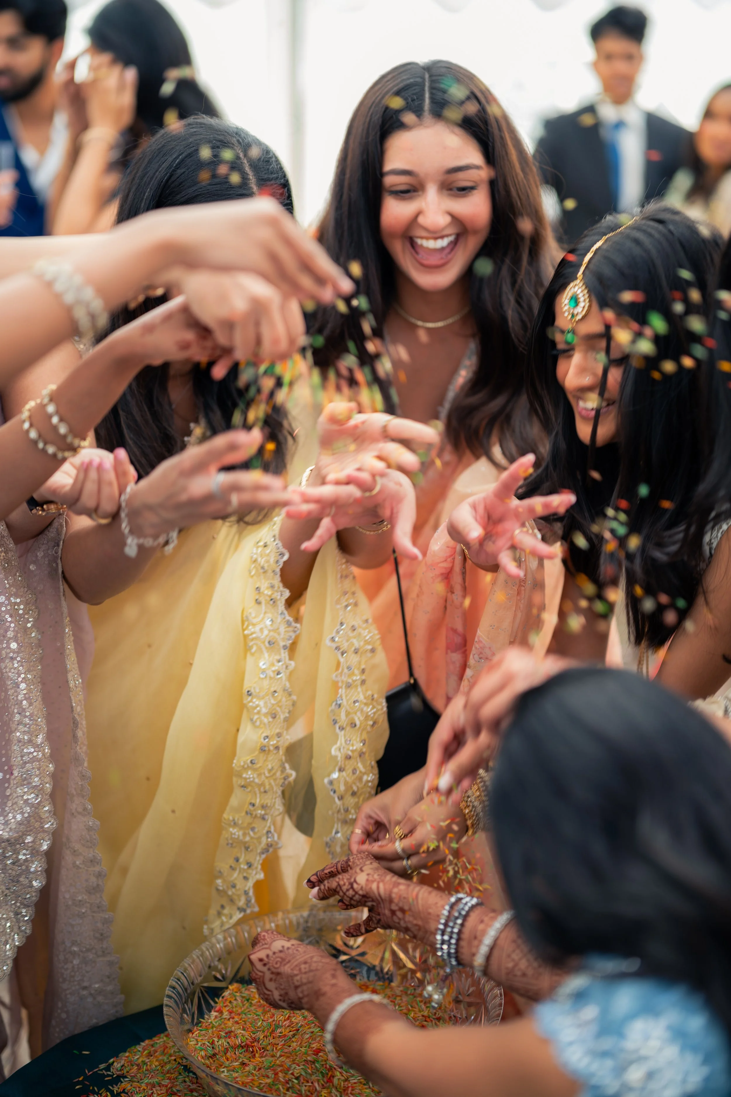 Women dressed in traditional Indian attire celebrating a festival with colorful rice and confetti at a lively gathering.