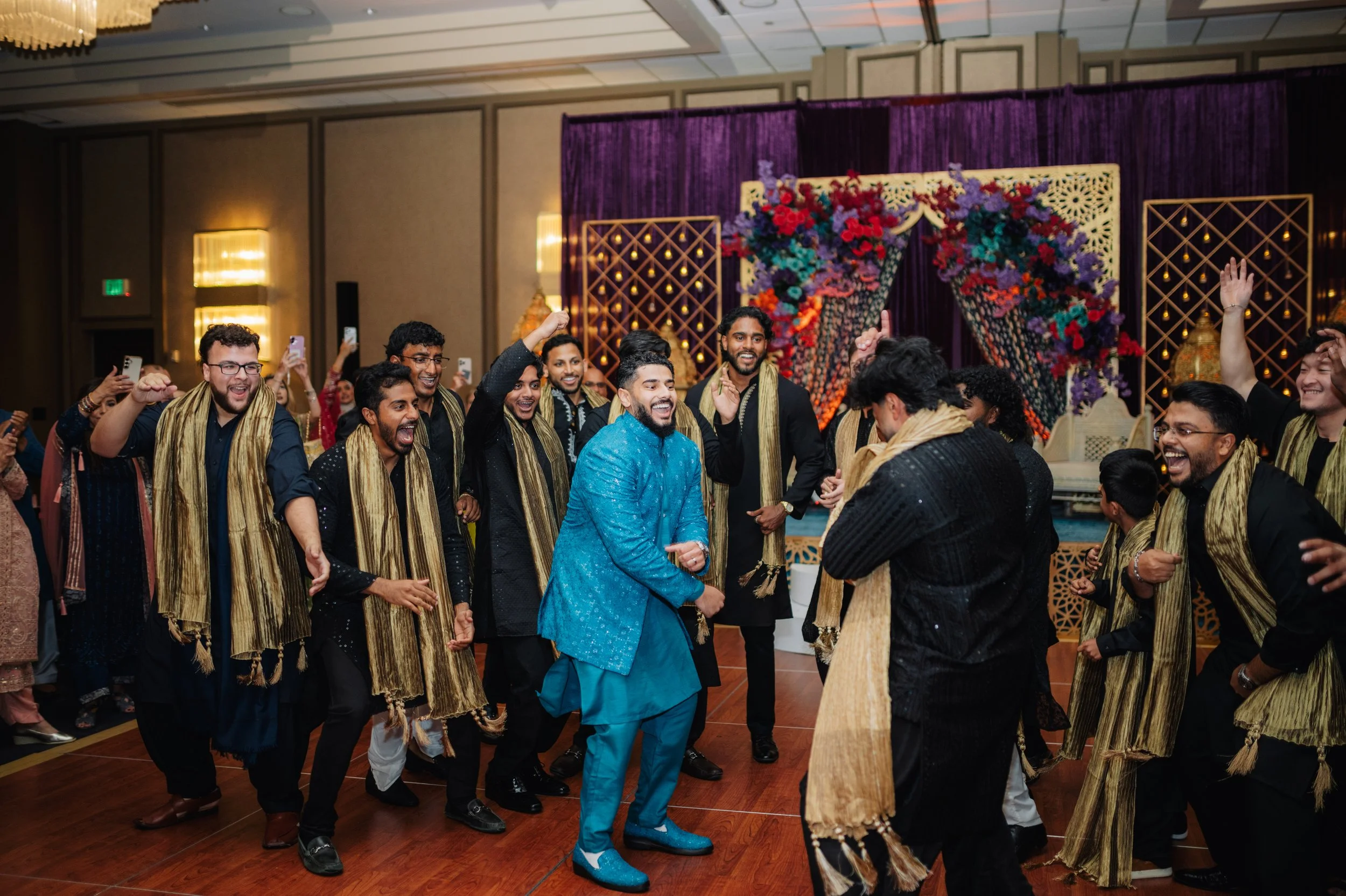 A group of people dressed in traditional Indian attire dancing and celebrating at a wedding or festive event, in a decorated hall with purple and gold decor.