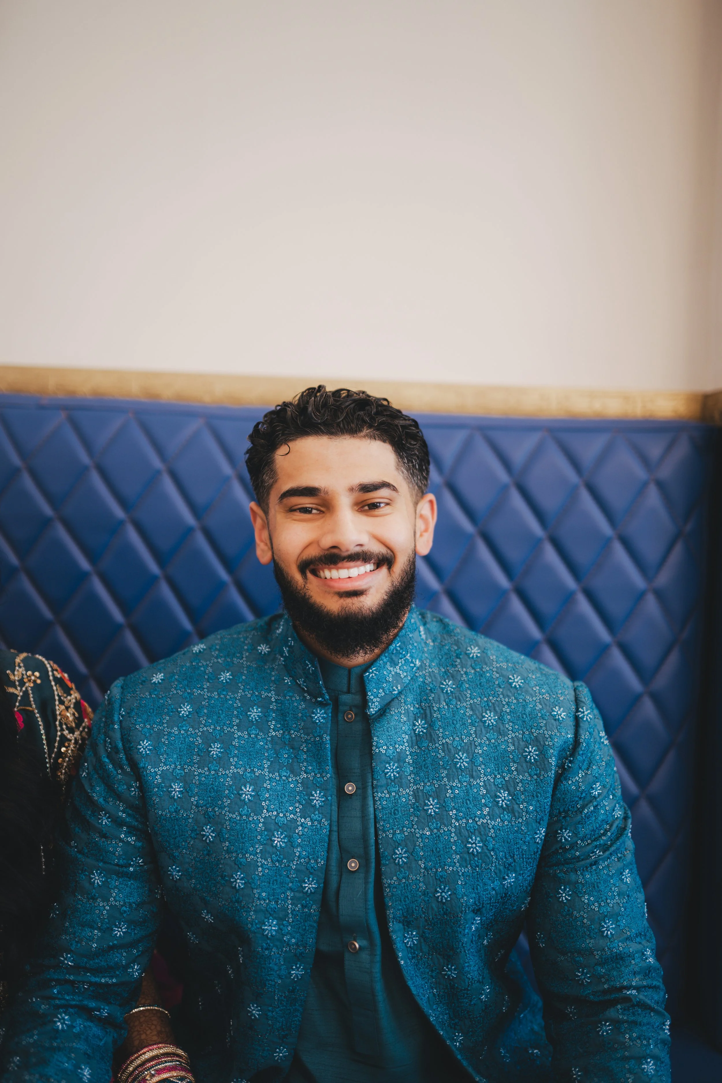 Portrait of a smiling man with dark hair and a beard, wearing a blue patterned traditional Indian jacket, sitting on a blue padded bench.