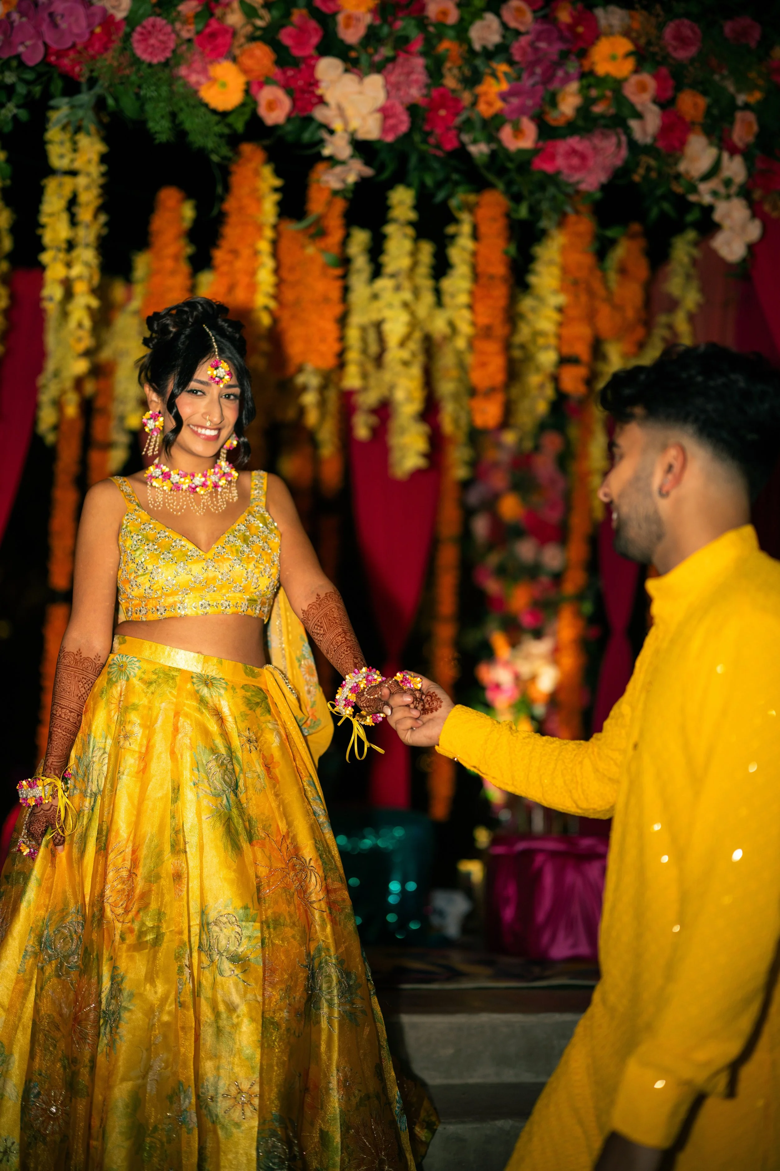 A woman and a man dressed in traditional Indian wedding attire, holding hands and smiling. The woman wears a yellow lehenga with floral embroidery, jewelry, and henna on her hands. The man wears a yellow kurta. The background is decorated with colorf
