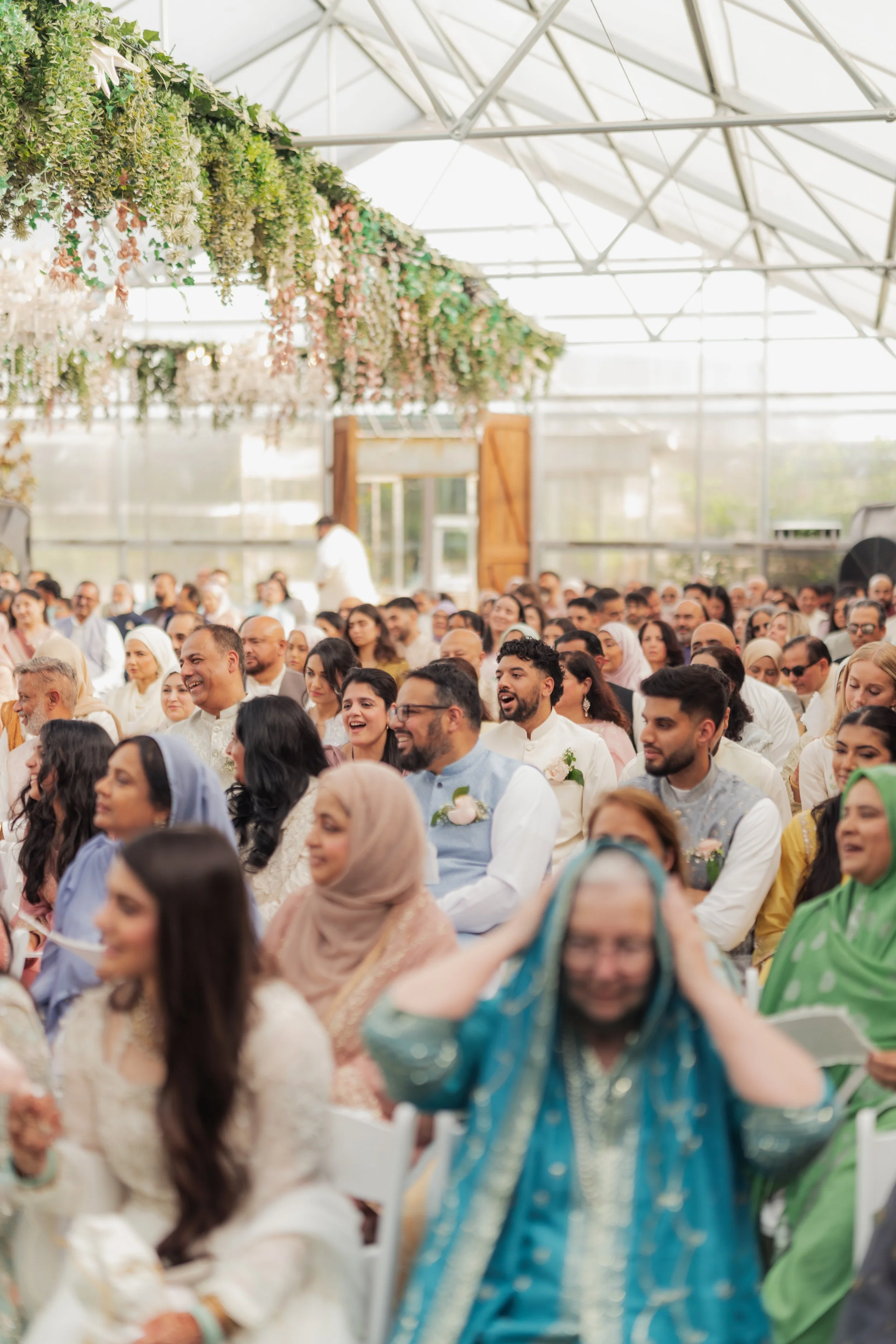 A large gathering of diverse people attending an indoor event in a greenhouse-like setting with glass walls, metal framing, decorated with hanging flowers and greenery.