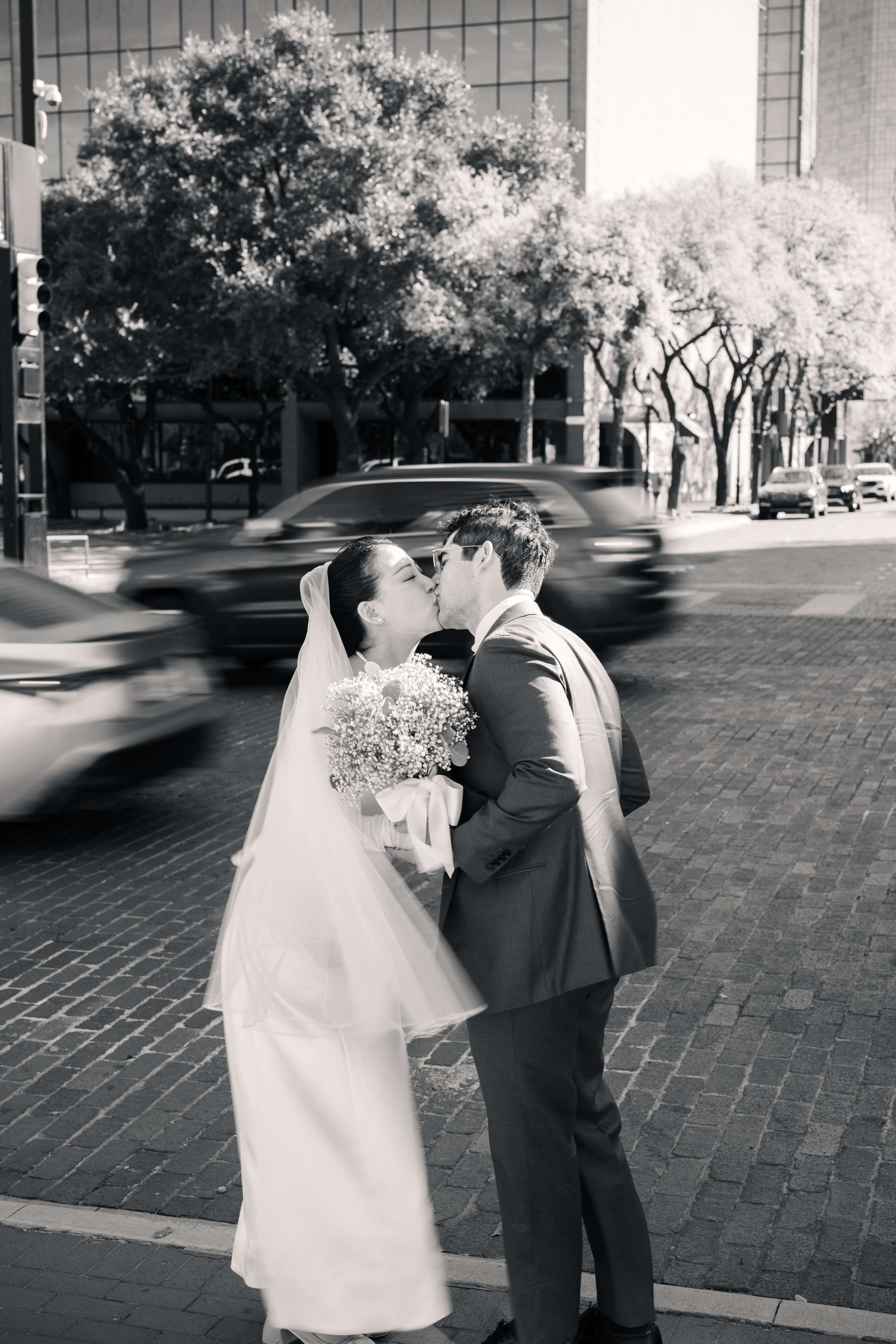A black-and-white photo of a bride and groom sharing a kiss on a city street, with blurred cars passing by in the background, and trees and modern buildings visible.