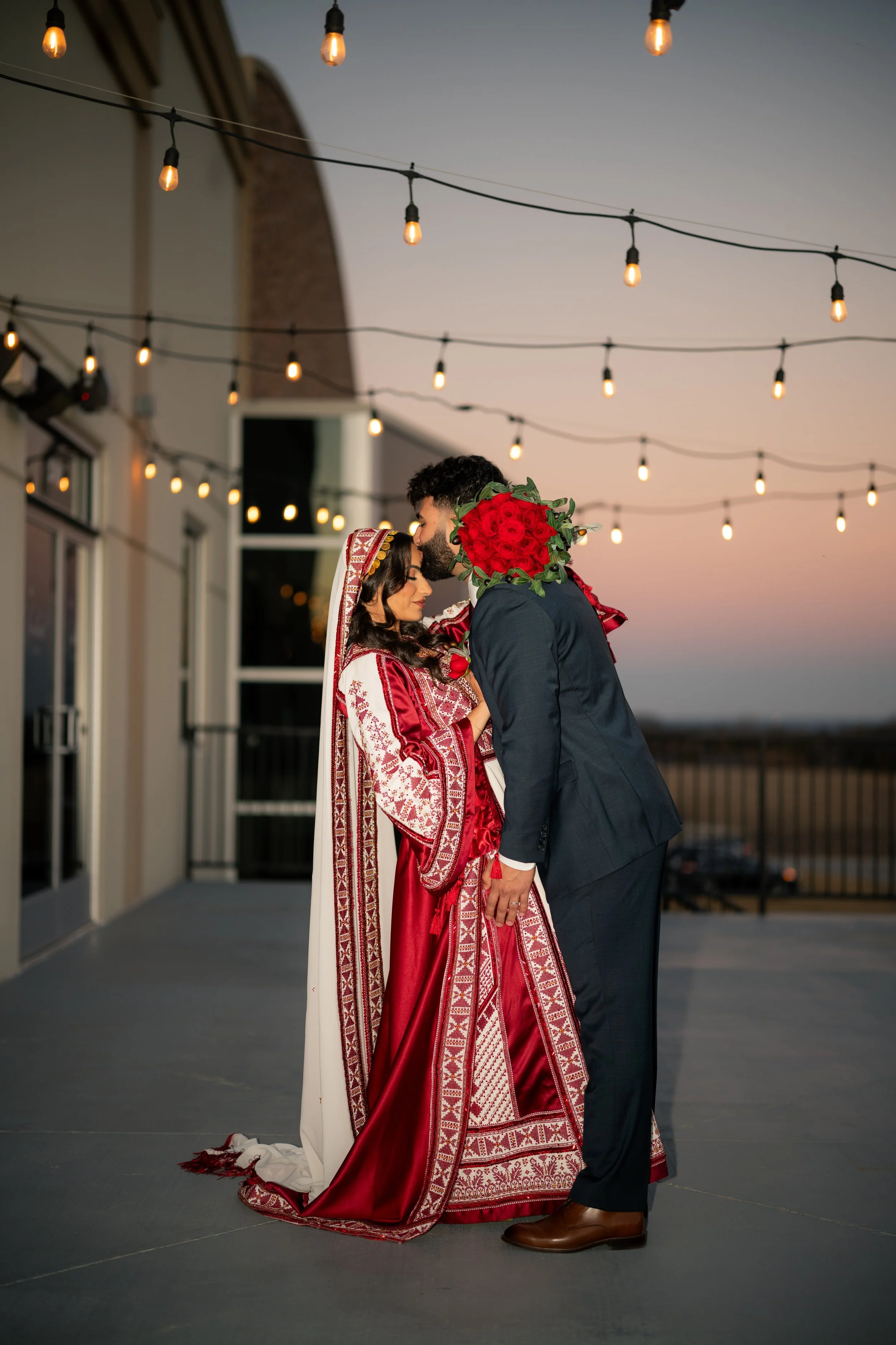 A couple dressed in traditional South Asian wedding attire, standing on a rooftop at sunset, with string lights overhead. The woman wears an intricate red and white embroidered dress with a veil, and the man wears a dark suit. The man holds a bouquet