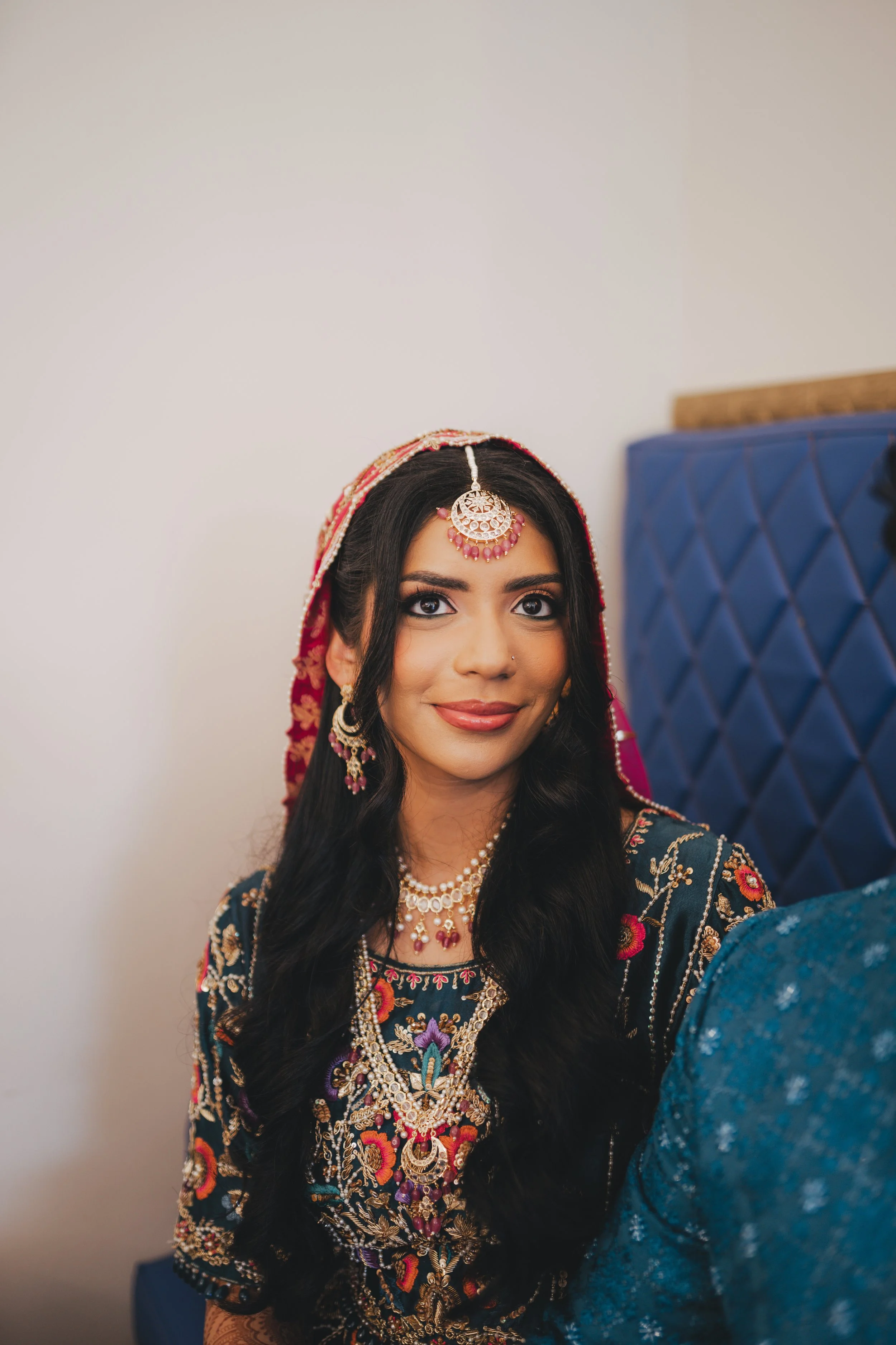 A woman with long black hair wearing traditional Indian jewelry and a colorful embroidered dress, smiling indoors.