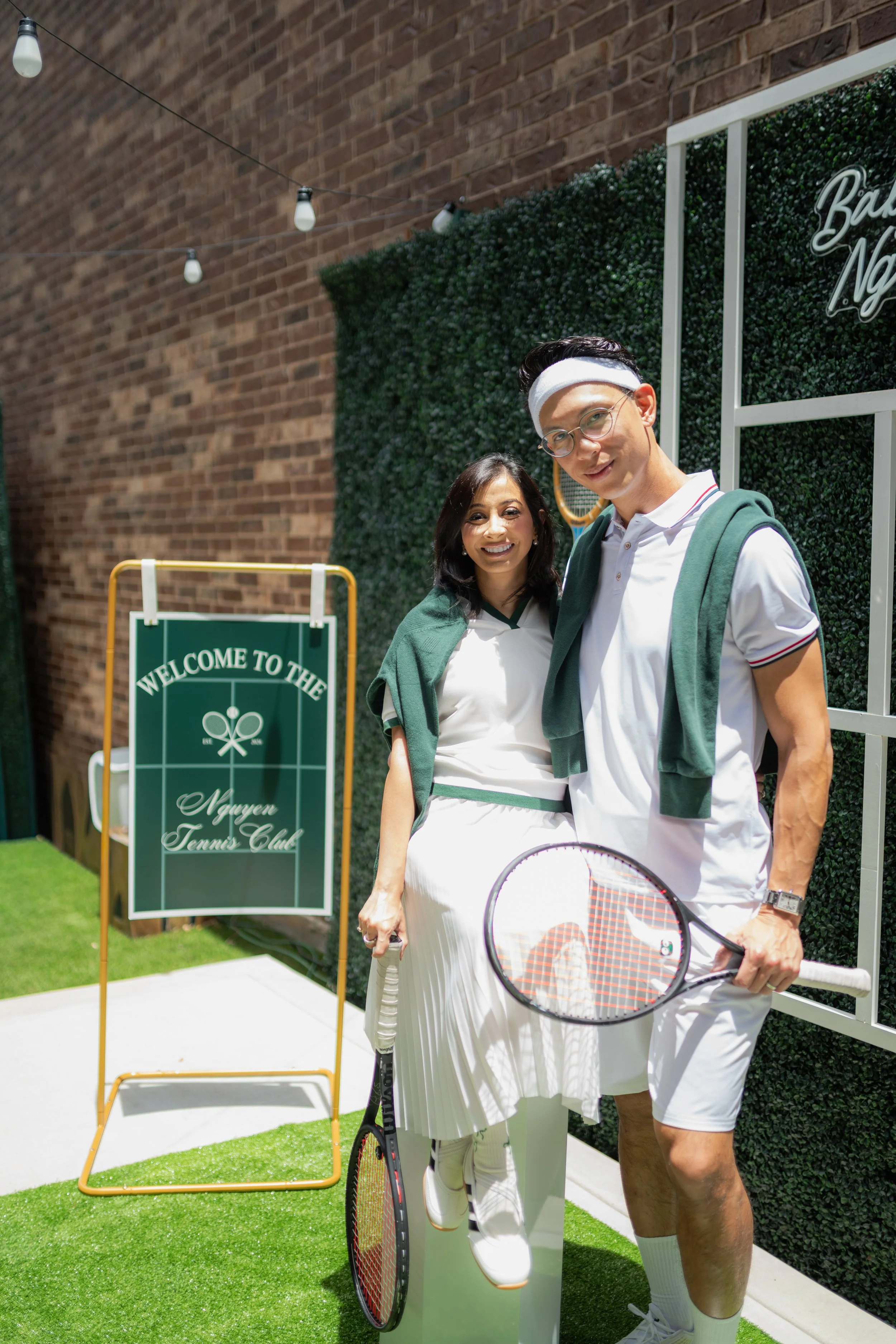 Two people, a woman and a man, dressed in tennis attire standing outdoors at a tennis club. The woman is holding a tennis racket, and both are smiling at the camera. There is a green sign reading "Welcome to the Nguyen Tennis Club" and a tennis net i