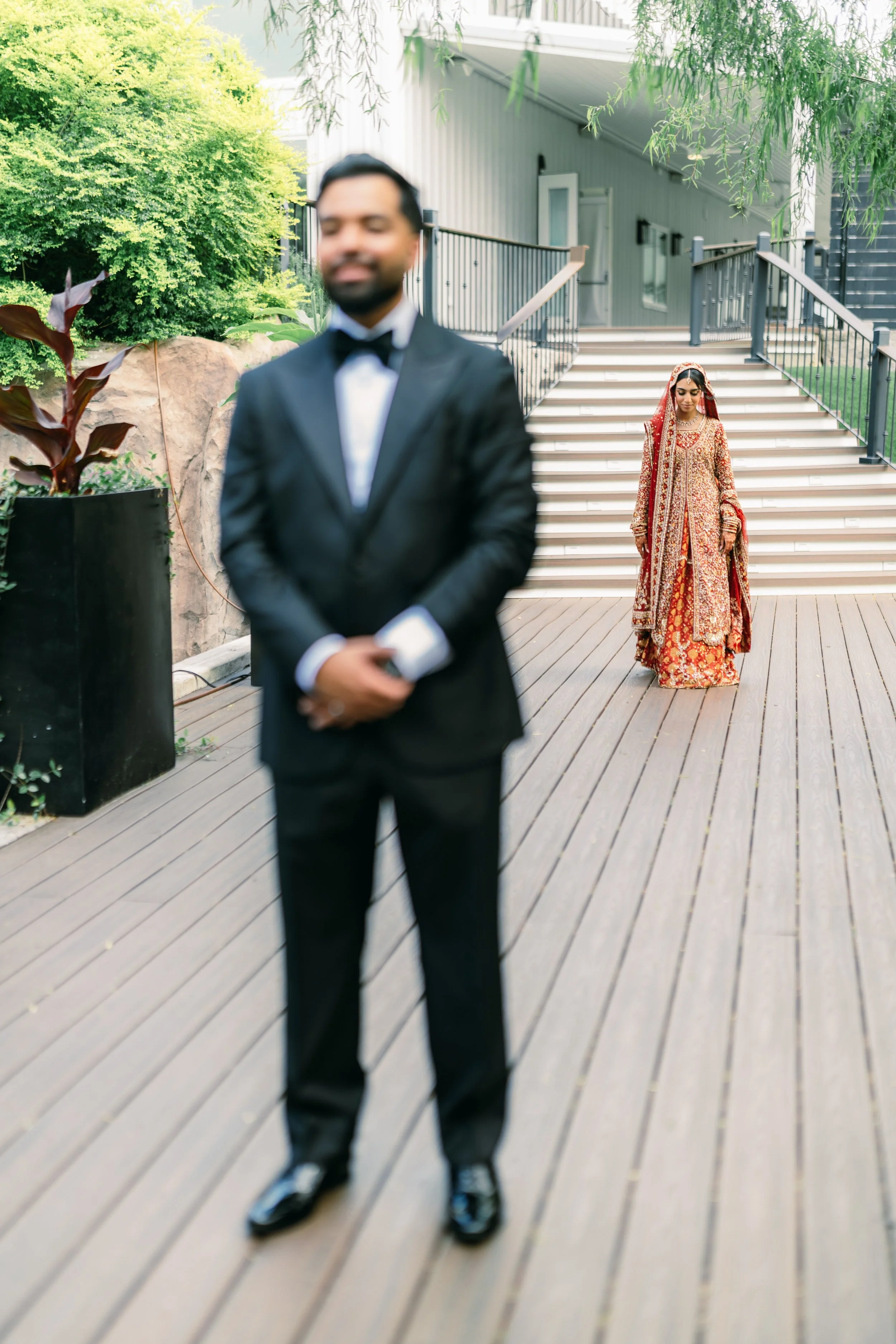 A man in a tuxedo with a bowtie standing on a wooden deck, with a woman in traditional red and gold bridal attire walking down stairs in the background.