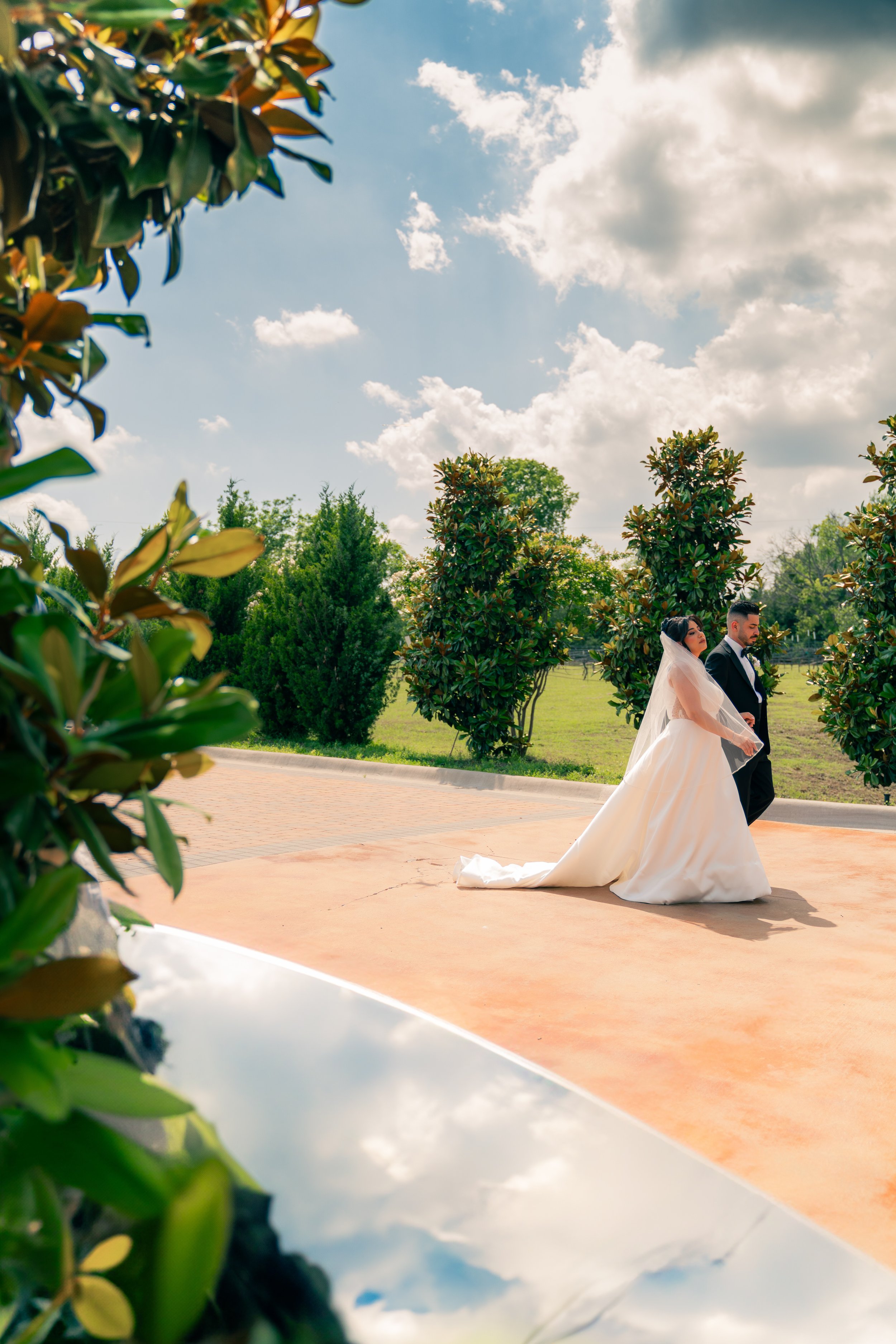 A bride in a white wedding dress and veil holding hands with a groom in a black tuxedo walking outdoors on a sunny day with a blue sky and clouds, surrounded by green trees.