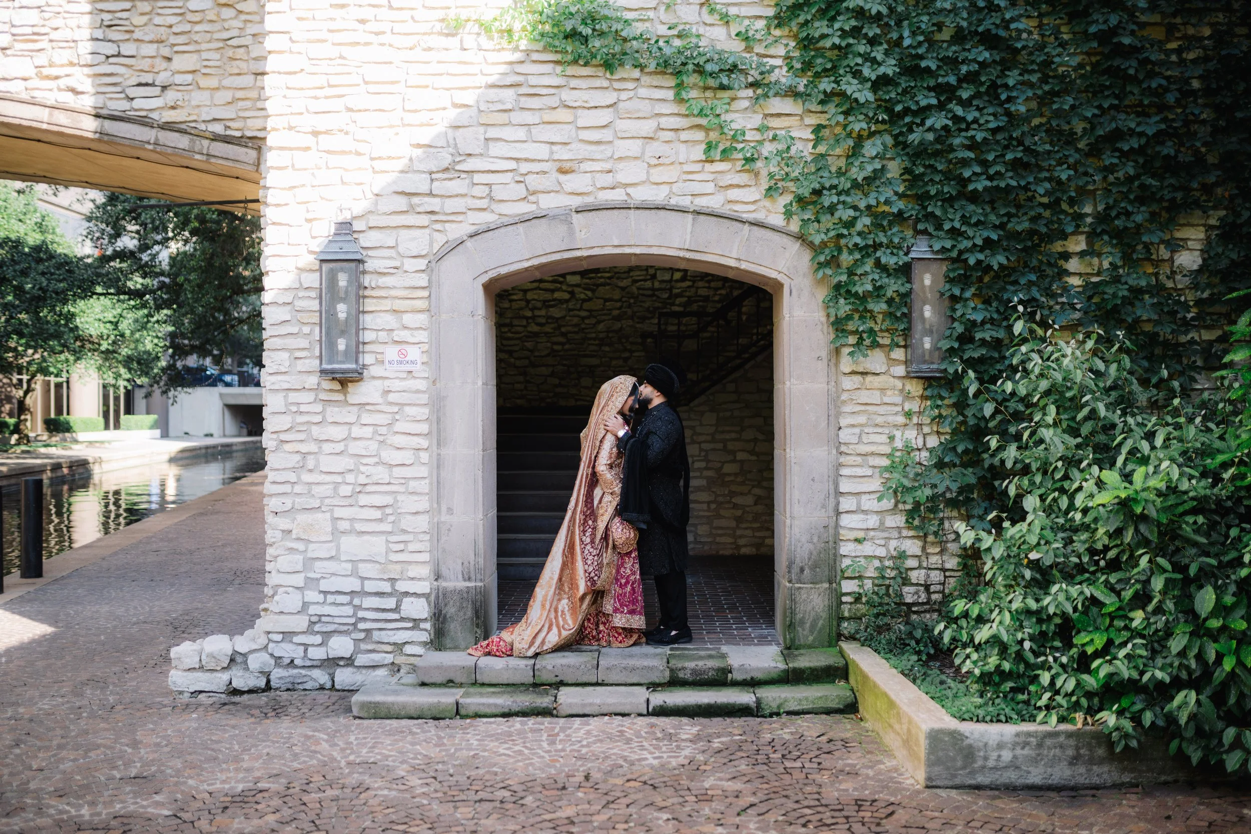 A couple dressed in traditional Indian wedding attire sharing a kiss under an archway with stone walls and greenery.