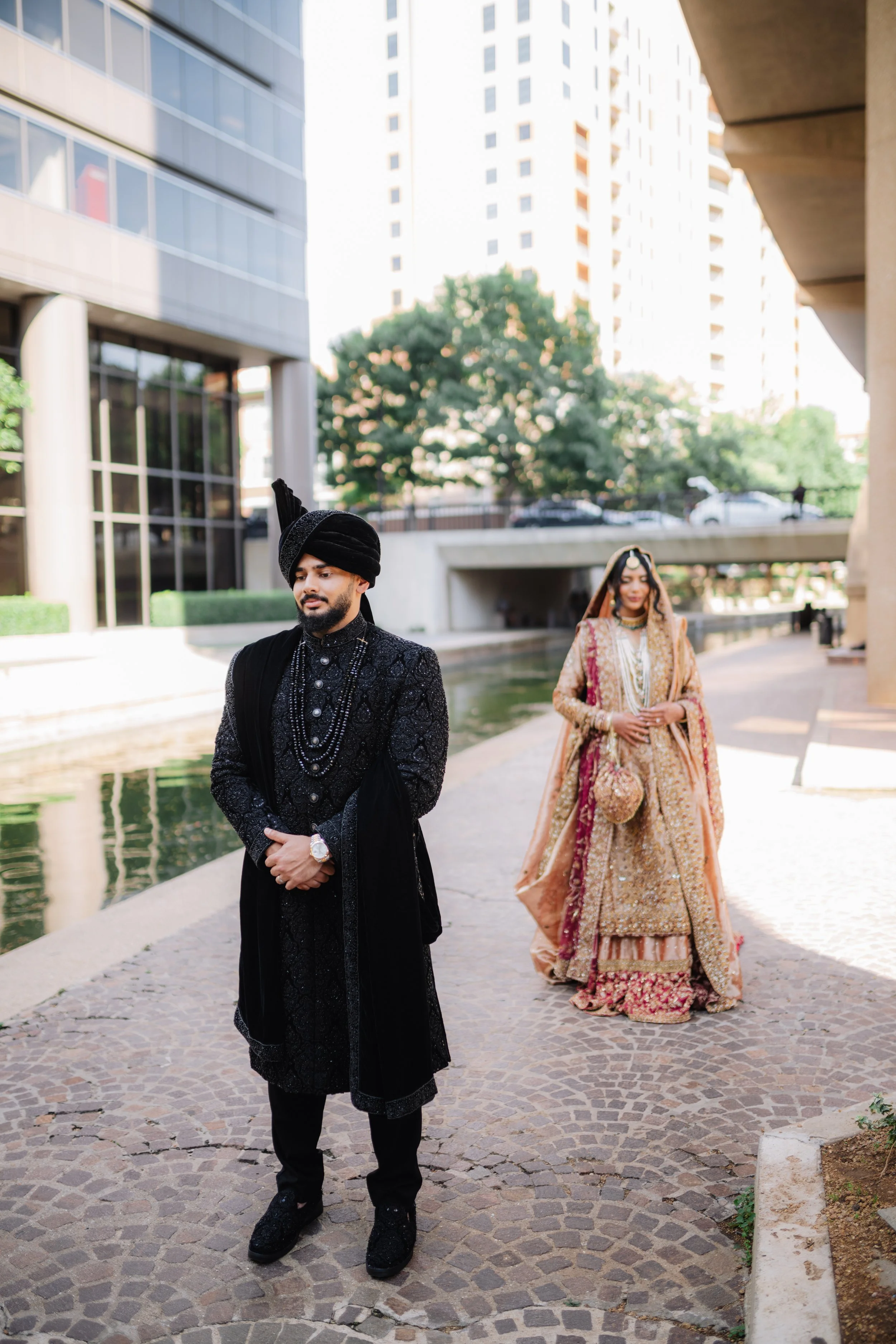 A man and woman dressed in traditional Indian wedding attire standing outdoors near a water feature with modern buildings in the background.