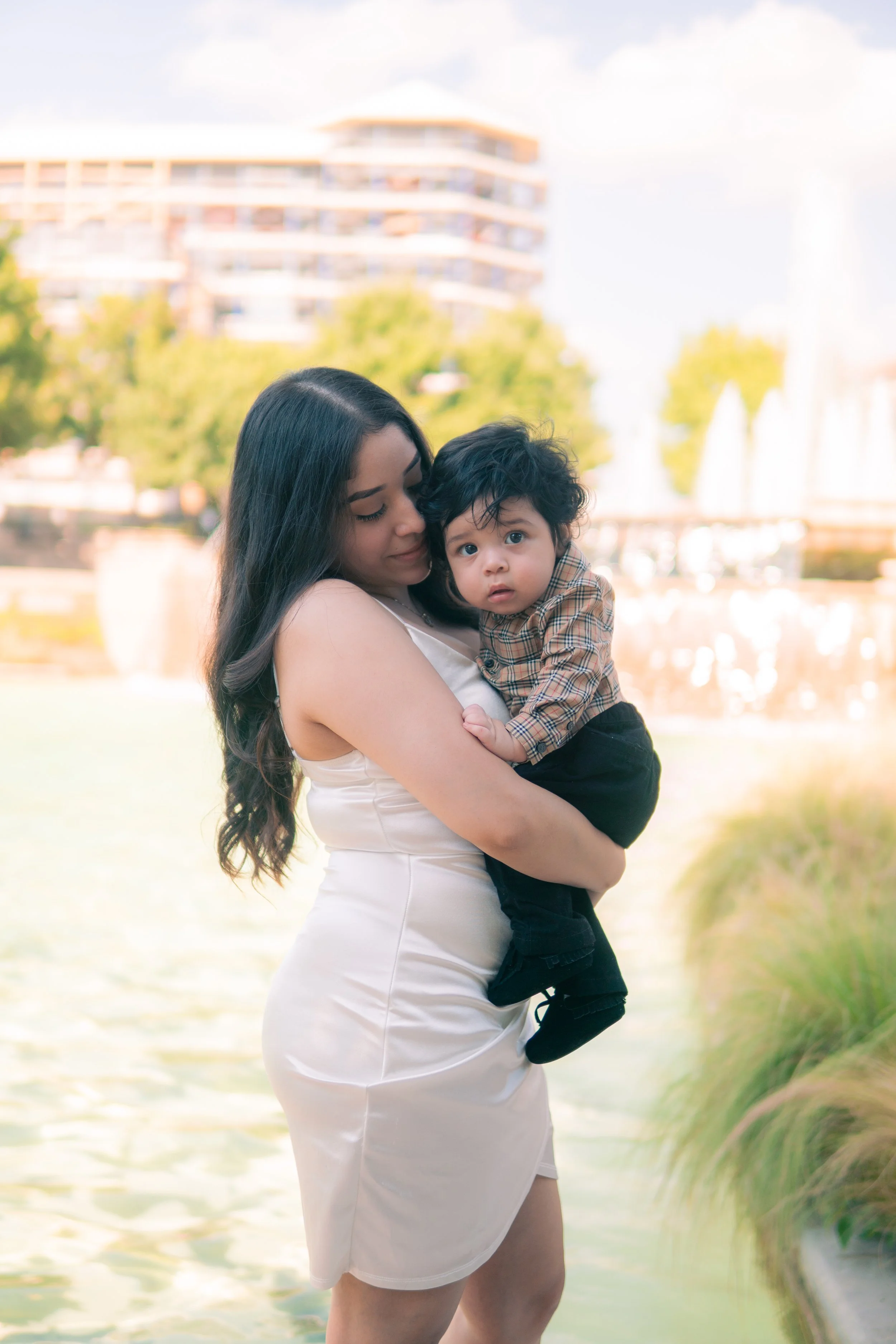 A woman holding a young boy outdoors by a water feature with buildings and trees in the background.