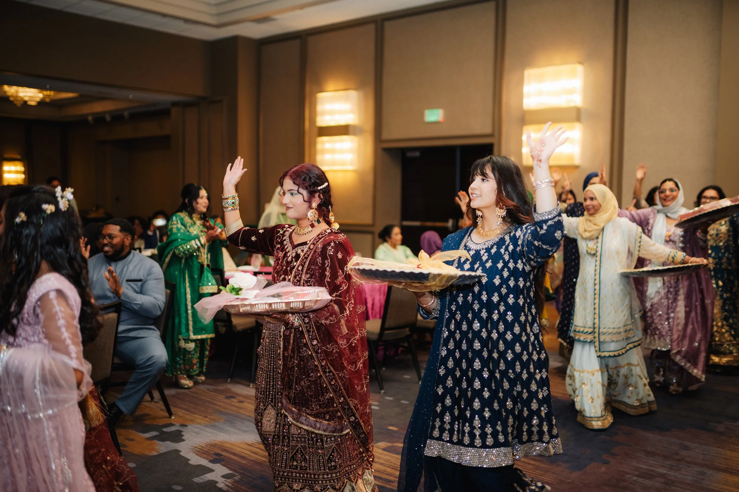 Women in traditional South Asian attire dancing at celebration event. One woman in blue holding a tray, others smiling and raising hands.