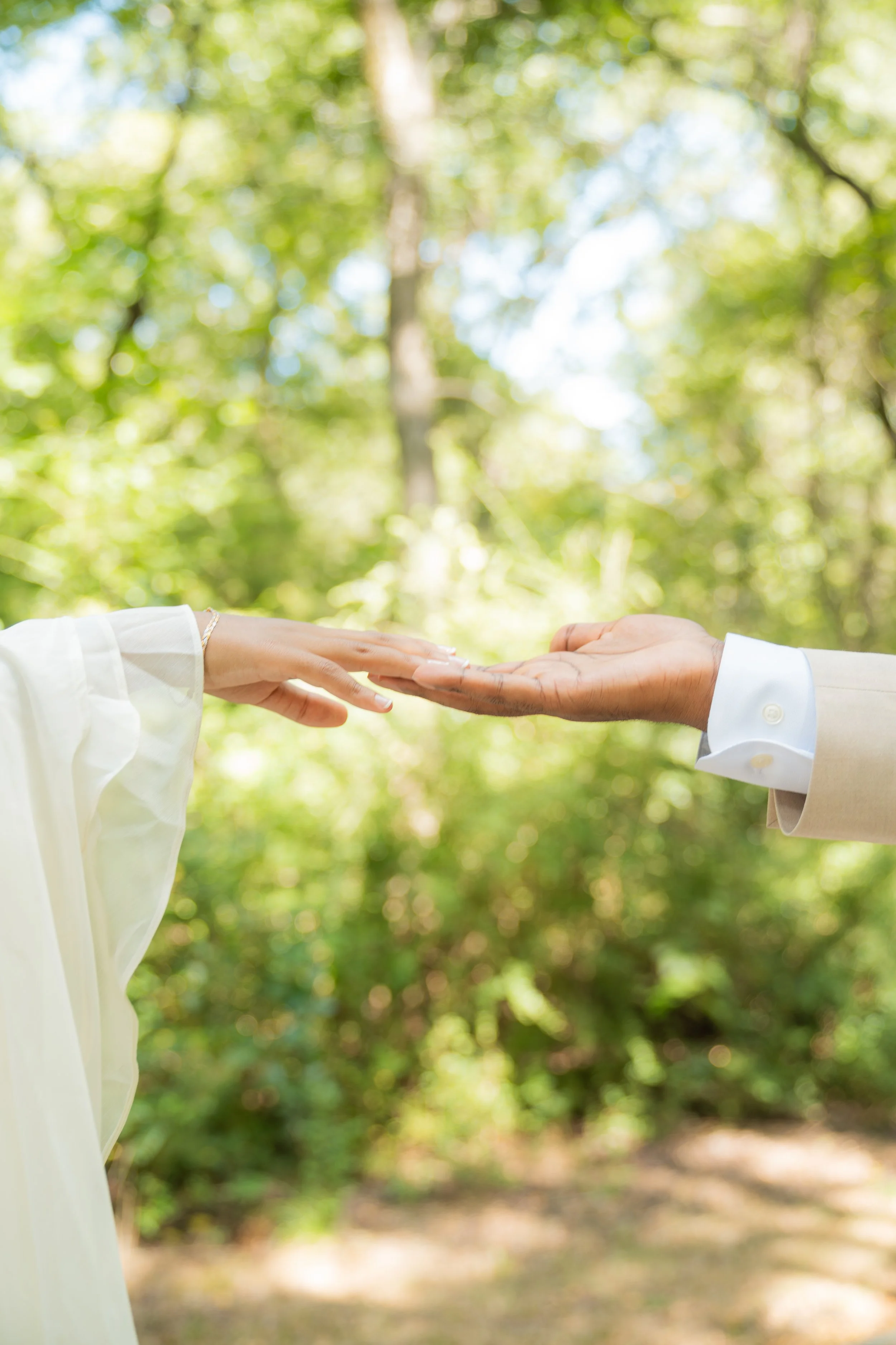 A couple holding hands with their fingers touching, outdoors in a lush green forest, dressed in formal attire.