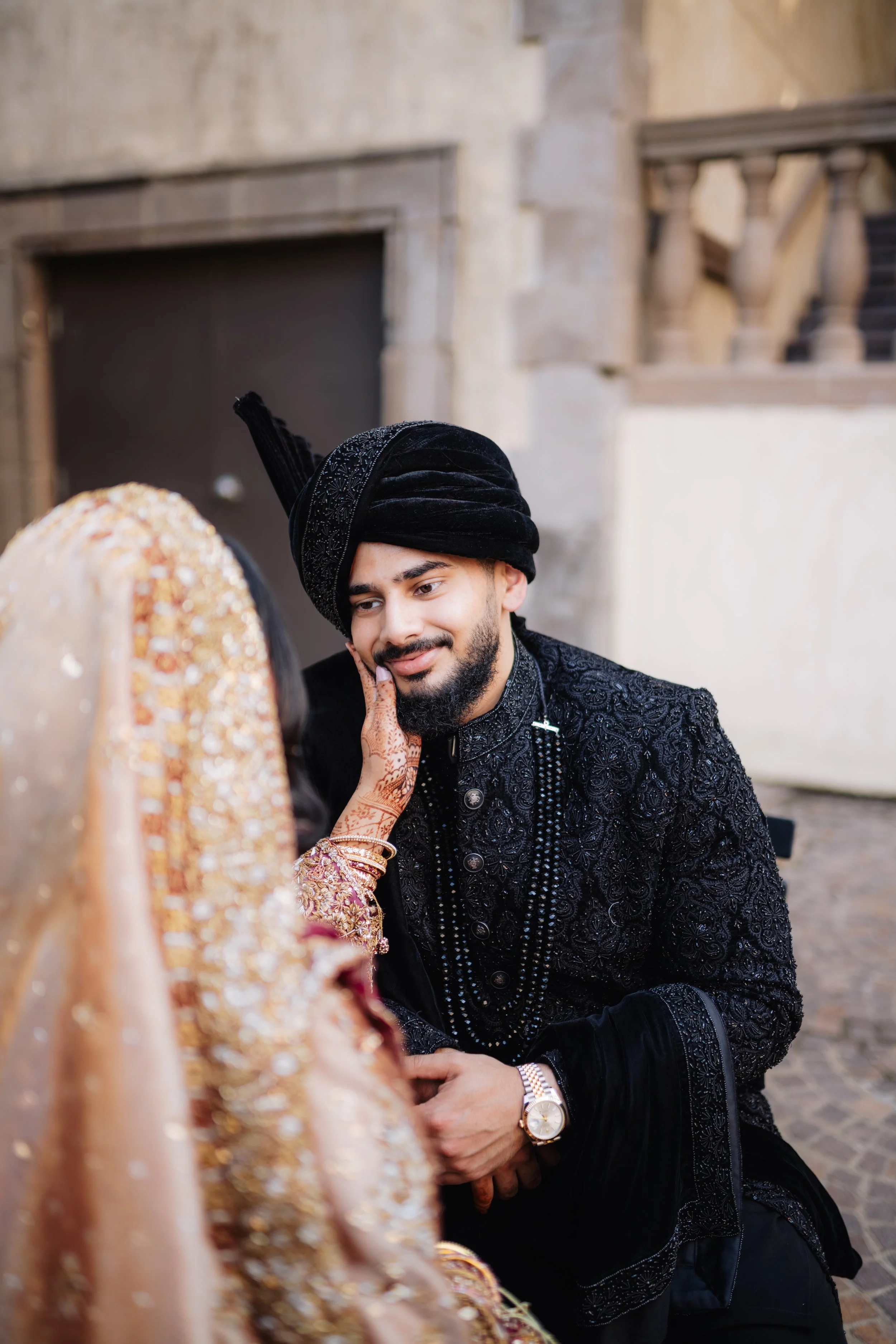 A man dressed in traditional South Asian attire with a black turban and ornate black sherwani, smiling and holding hands with a woman in bright gold and pink attire, during a wedding ceremony.