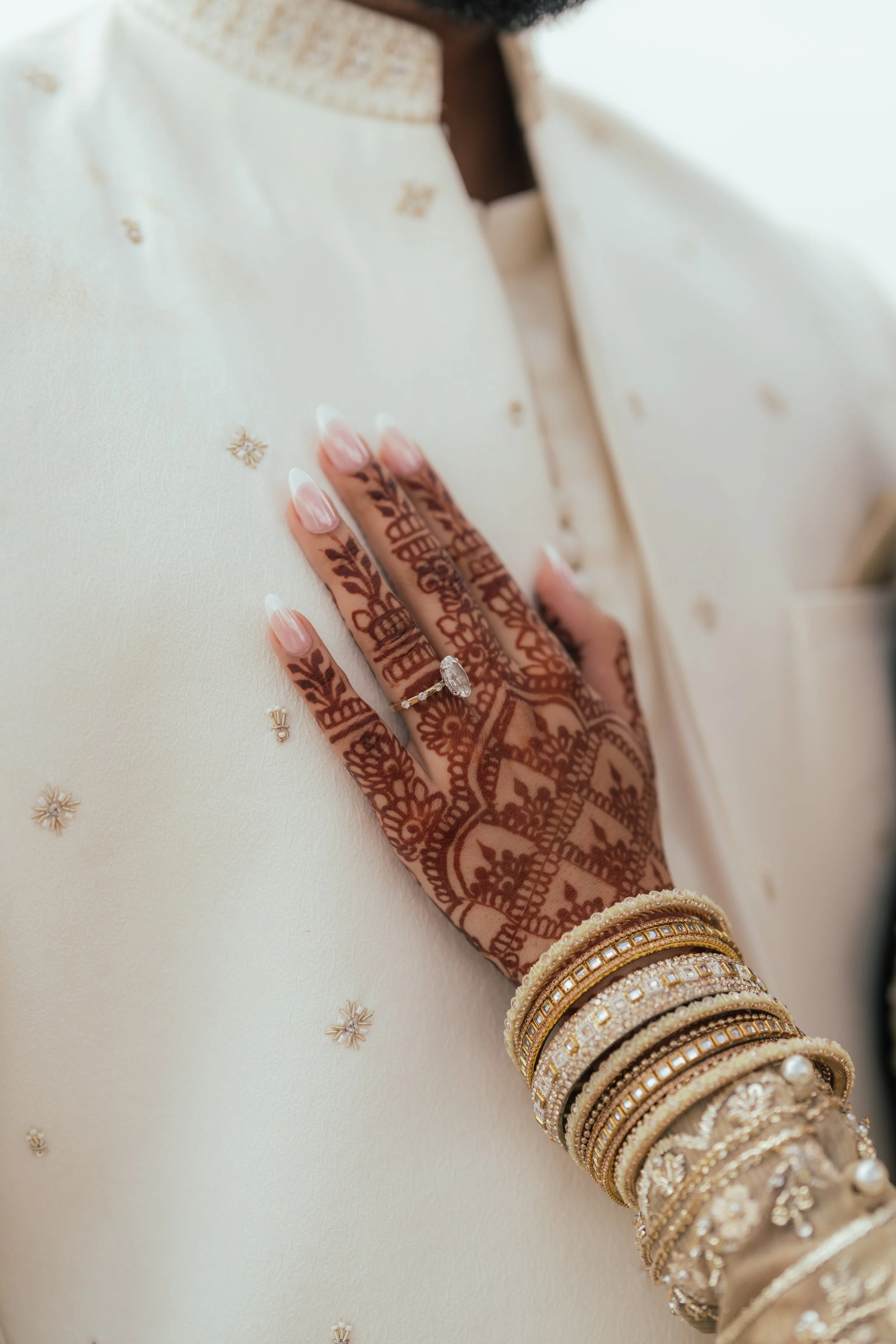 Close-up of a hand with henna designs, rings, and bangles placed on cream-colored fabric with small gold or pearl decorative accents.