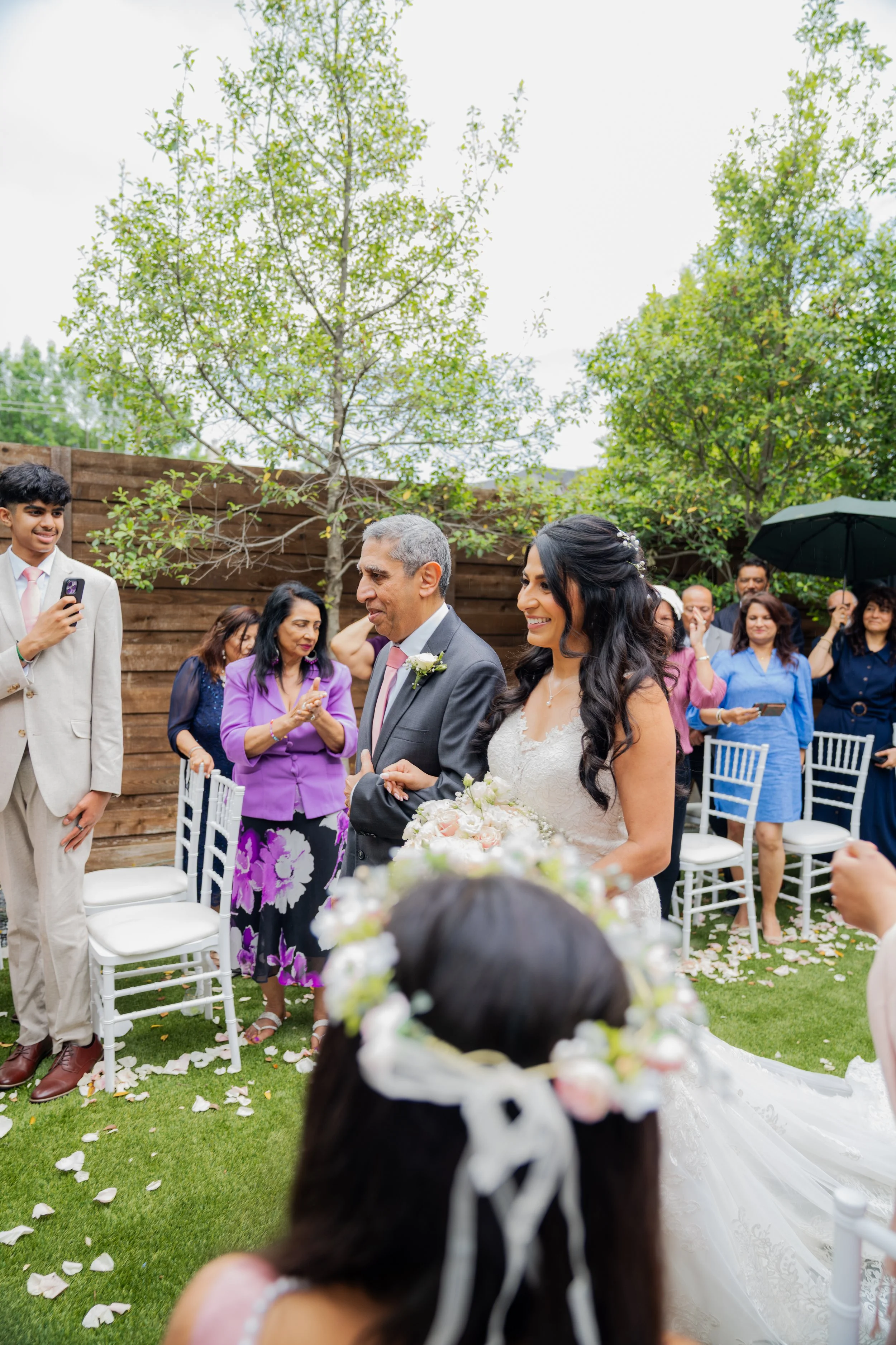 A wedding ceremony outdoors with a bride and groom exchanging vows, surrounded by friends and family. The bride is smiling, holding a bouquet, with flower petals on the grass. Guests are seated and standing, some taking photos, with trees and a woode