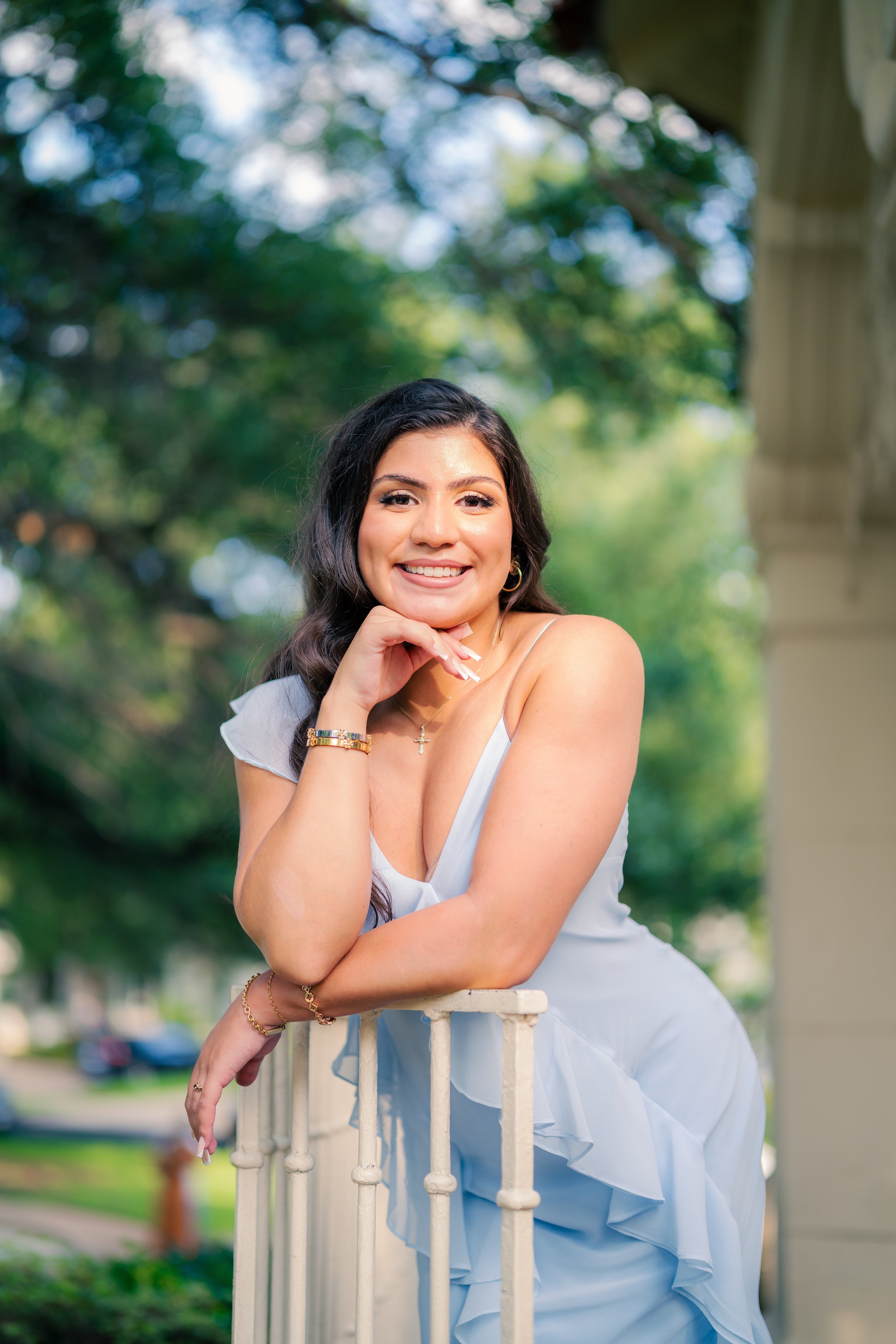 A smiling woman in a white dress leaning on a railing outdoors with green trees in the background.