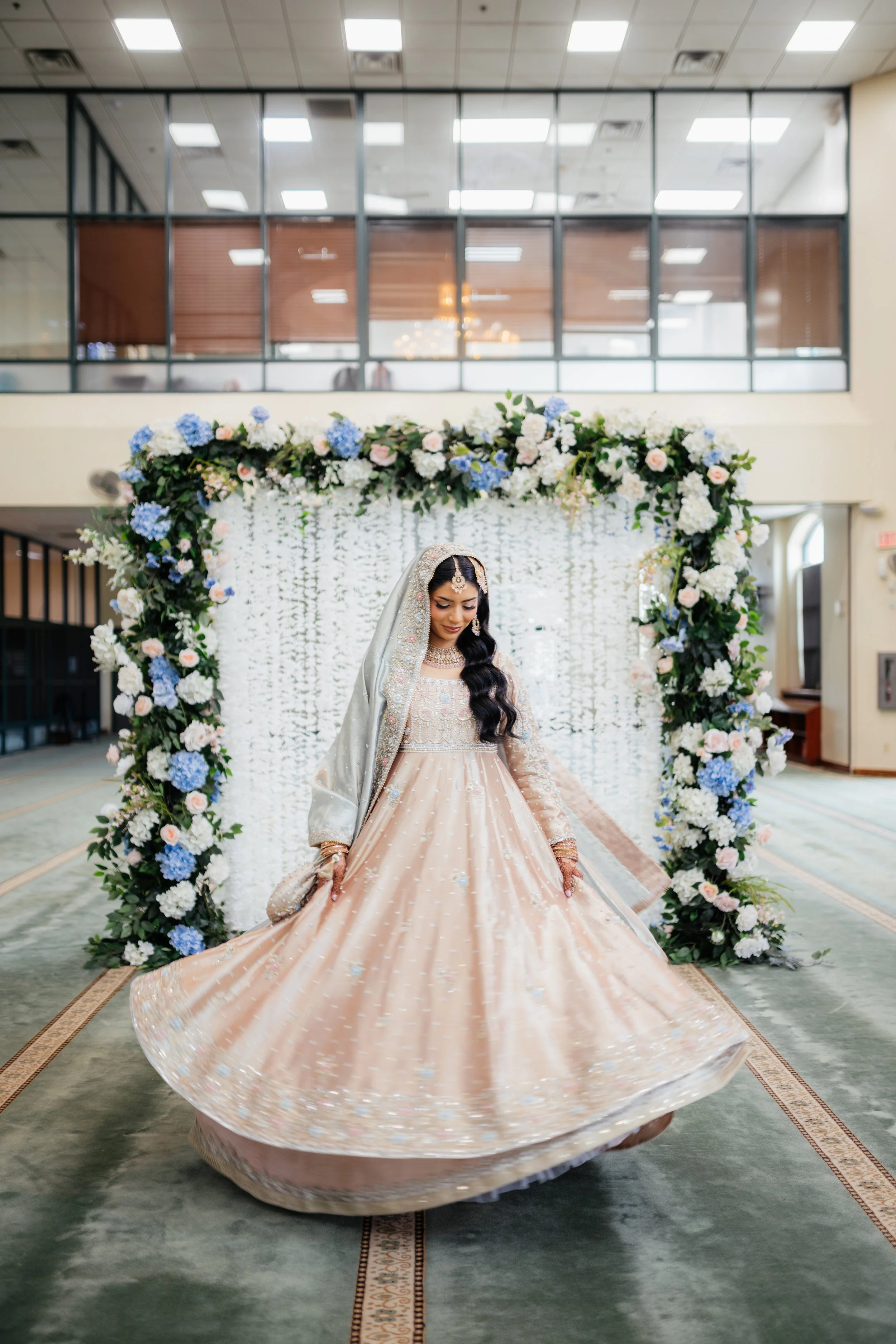 A bride in traditional South Asian attire twirling under a floral arch at her wedding ceremony inside a spacious hall.