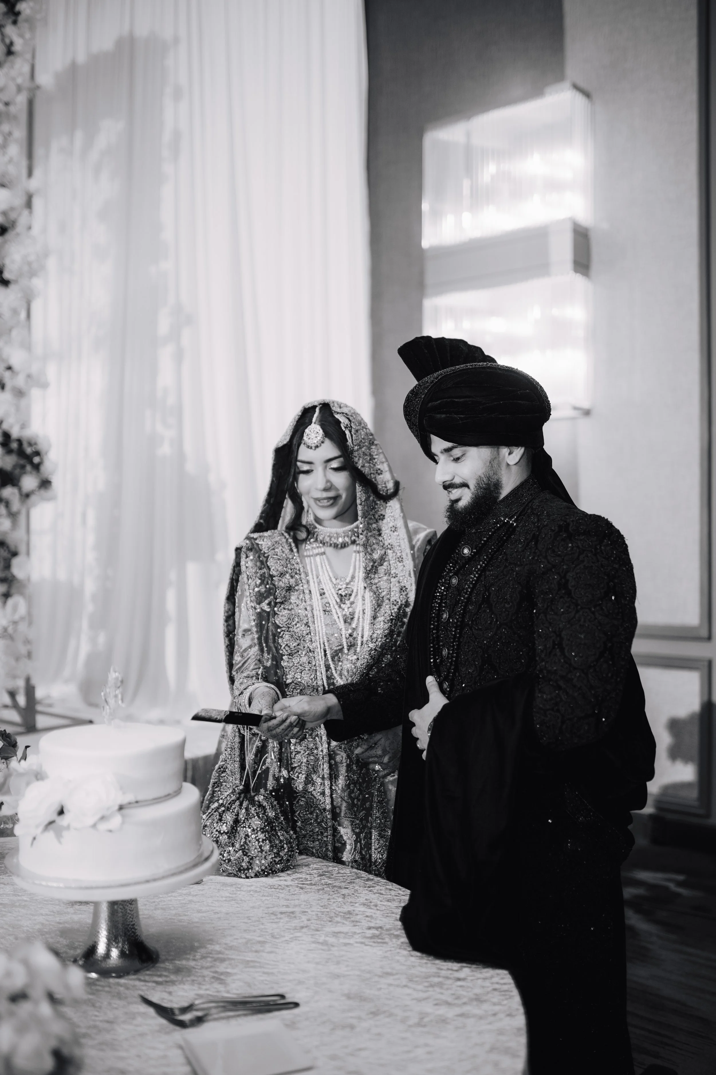 A bride and groom in traditional South Asian wedding attire at a wedding reception, standing next to a wedding cake, looking at a phone or a card together.