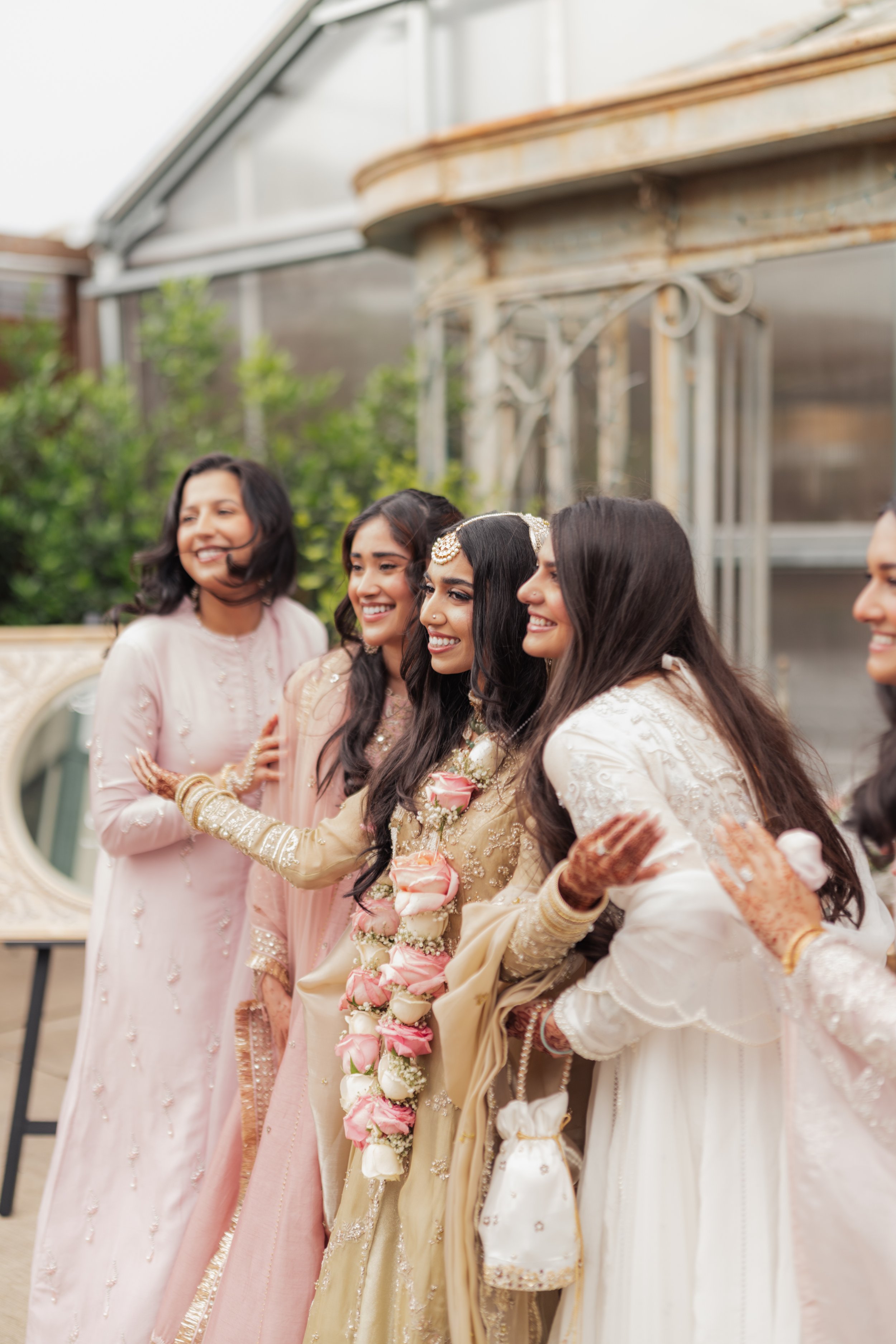 A group of women dressed in traditional South Asian attire at a celebration, smiling and holding hands, with a green garden in the background.