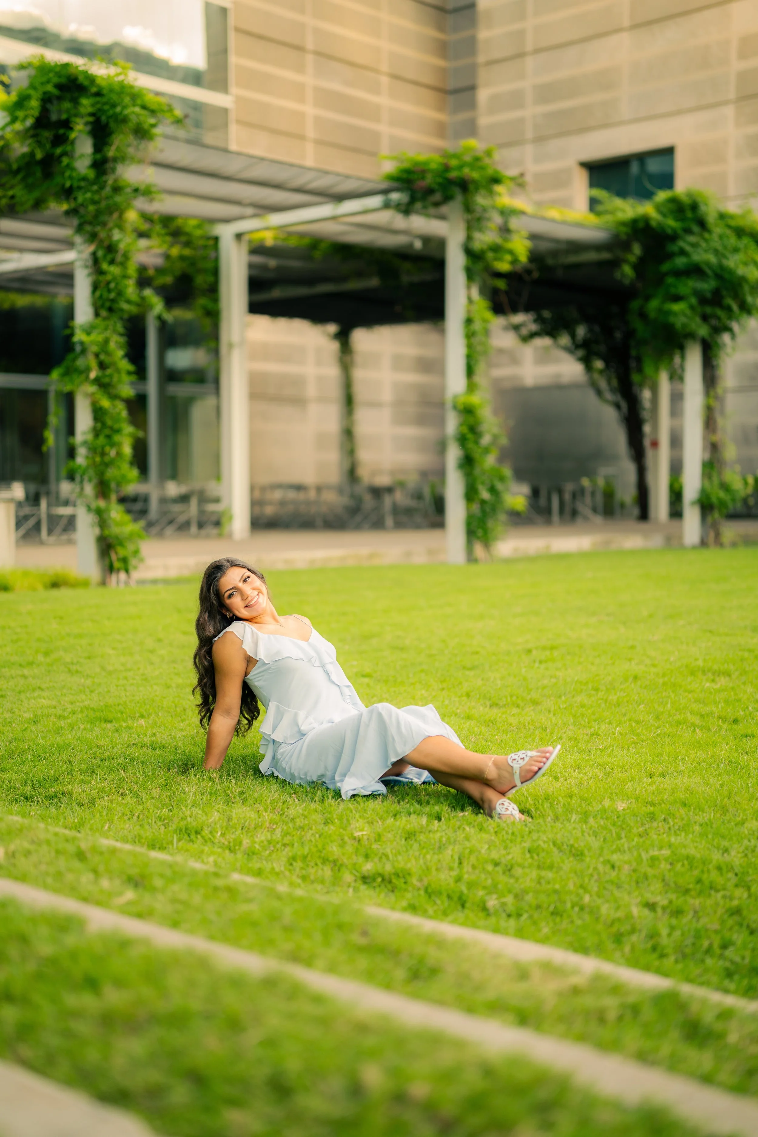 A woman in a white dress smiling and relaxing on a grassy lawn with trees and modern buildings in the background.
