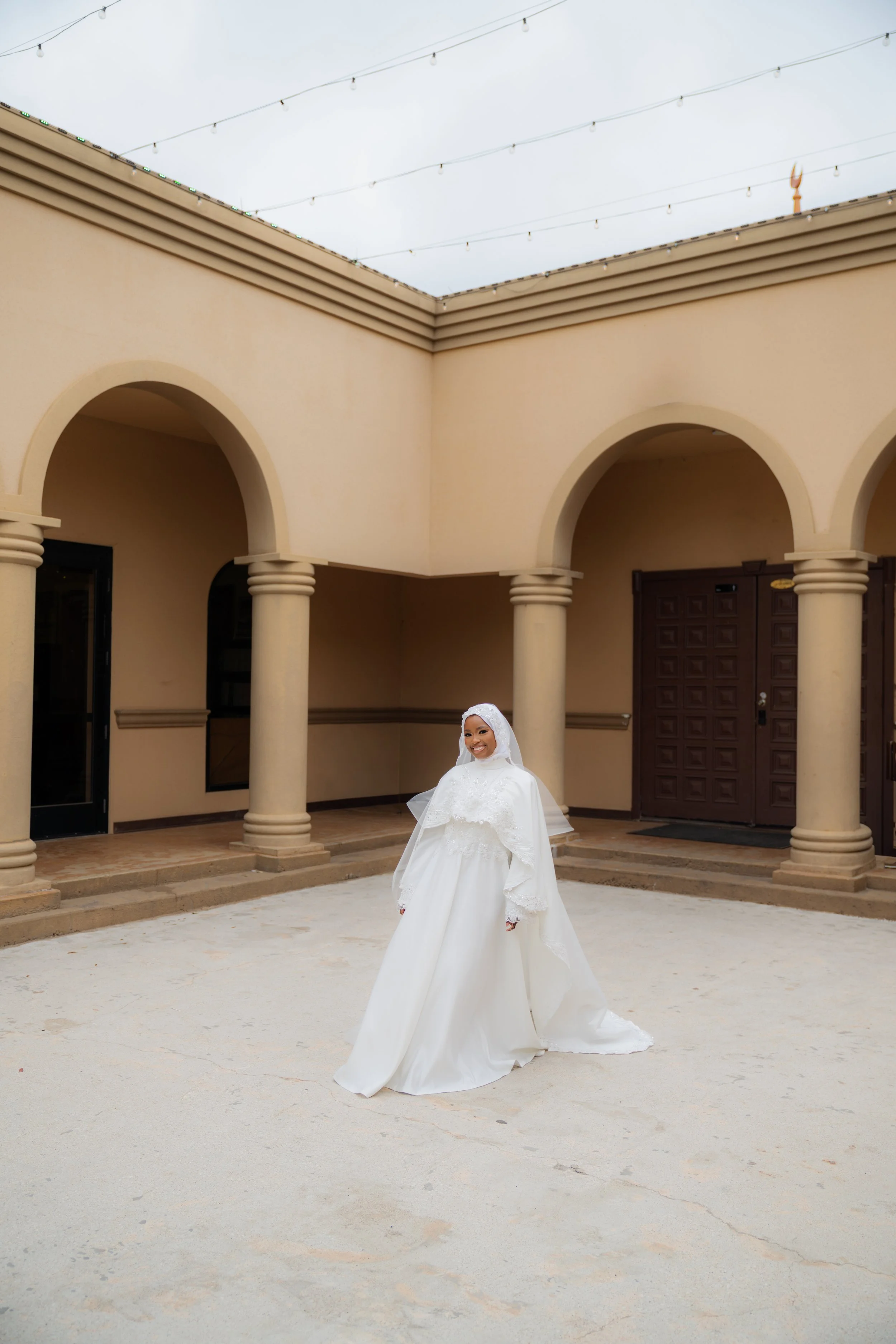 A bride in a white wedding dress and hijab standing in a courtyard with beige walls, arched doorways, and string lights above.