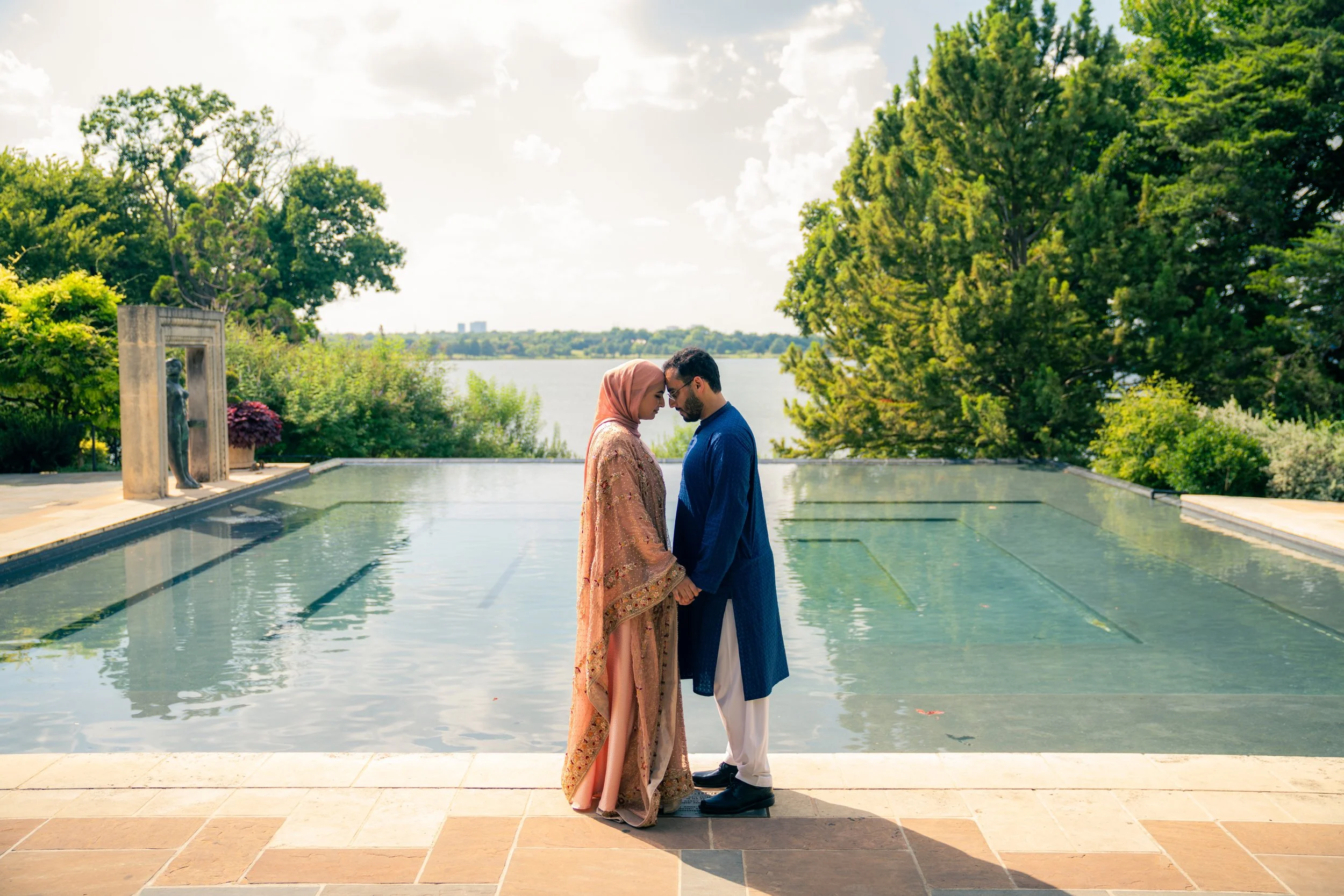 A couple dressed in traditional attire holding hands and leaning their foreheads together by a poolside with trees and water in the background.