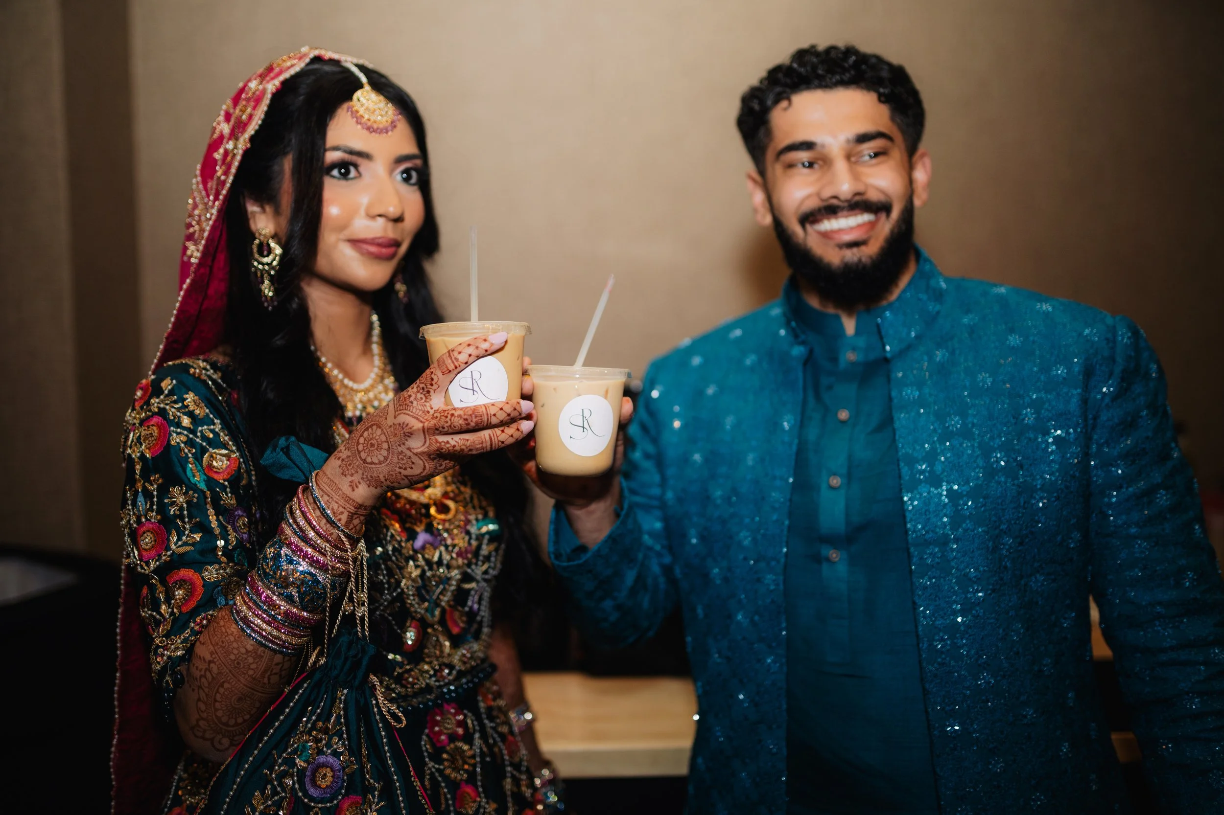 A woman and man dressed in traditional Indian attire, holding cups of mango lassi, smiling at an event.