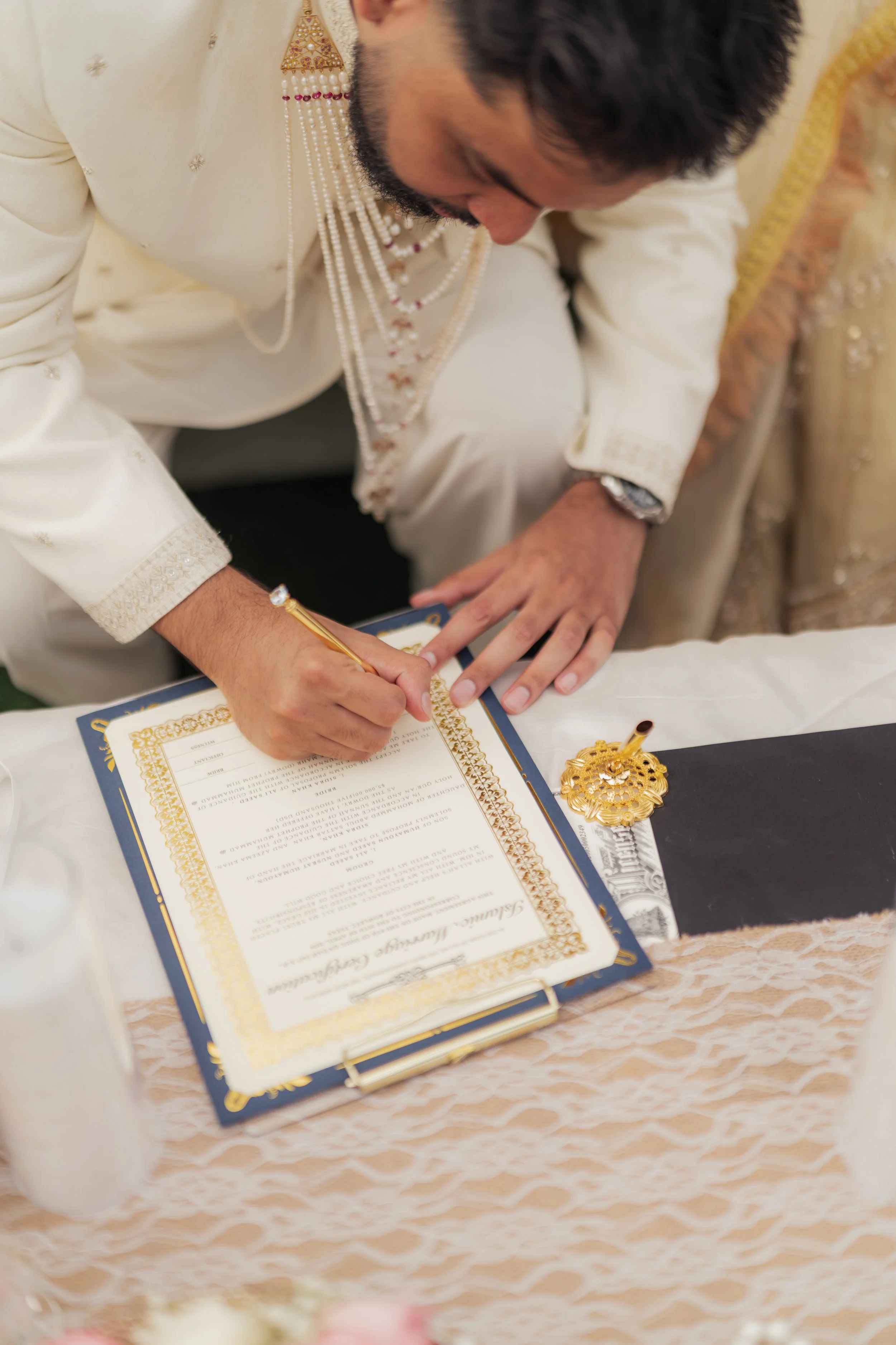 A man dressed in traditional formal attire signing a document at a table decorated with a lace tablecloth and gold embellishments.